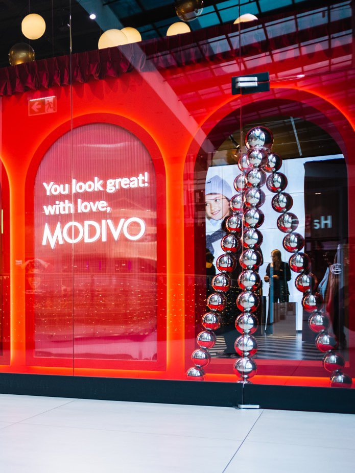 Store window display with red neon lighting, featuring a sign that says, 'You look great! with love, MODIVO.' There are decorative hanging metallic spheres and a large digital screen showing a smiling woman wearing a winter hat inside the store.