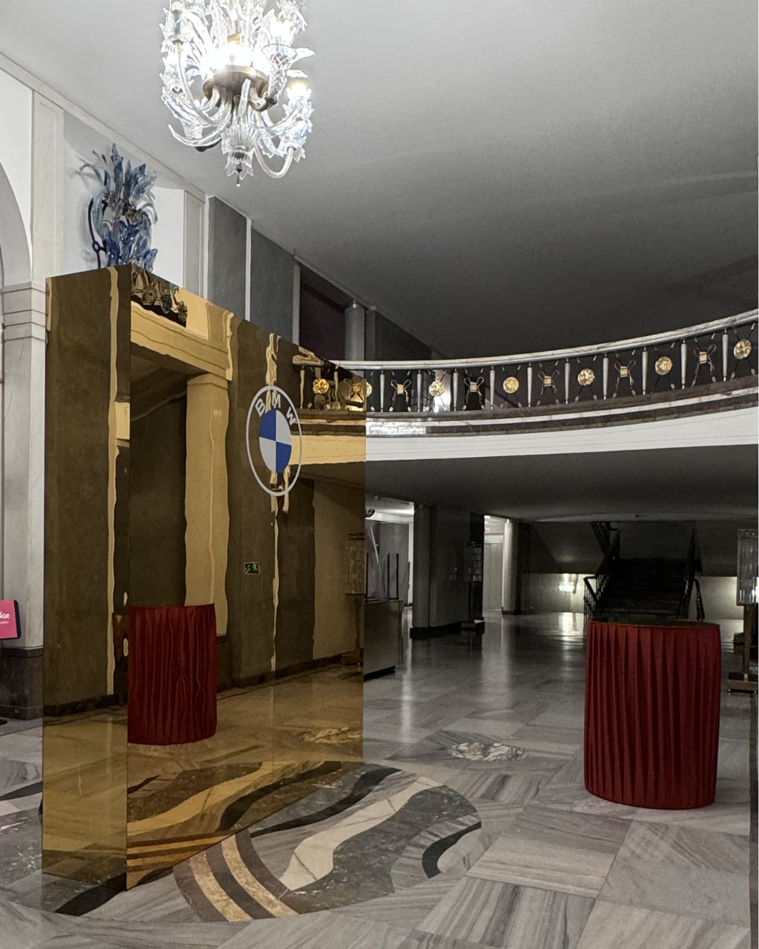 An interior of a building lobby with a custom mirror with a BMW logo, a chandelier, and a staircase. The mirror reflects part of the lobby with marble flooring.