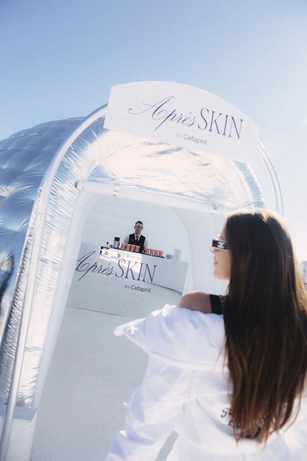 A woman with long dark hair and sunglasses standing in front of a white skincare booth labeled 'Aprés SKIN by Cetaphil' at an outdoor event on a sunny day.