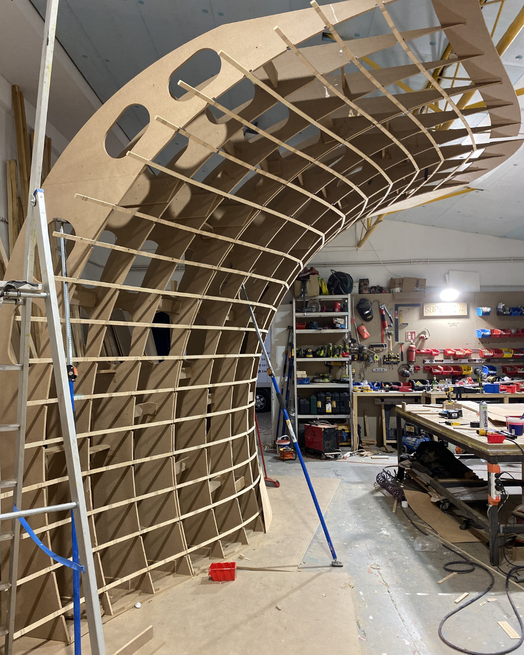 Wooden boat hull under construction in a workshop surrounded by tools and materials.