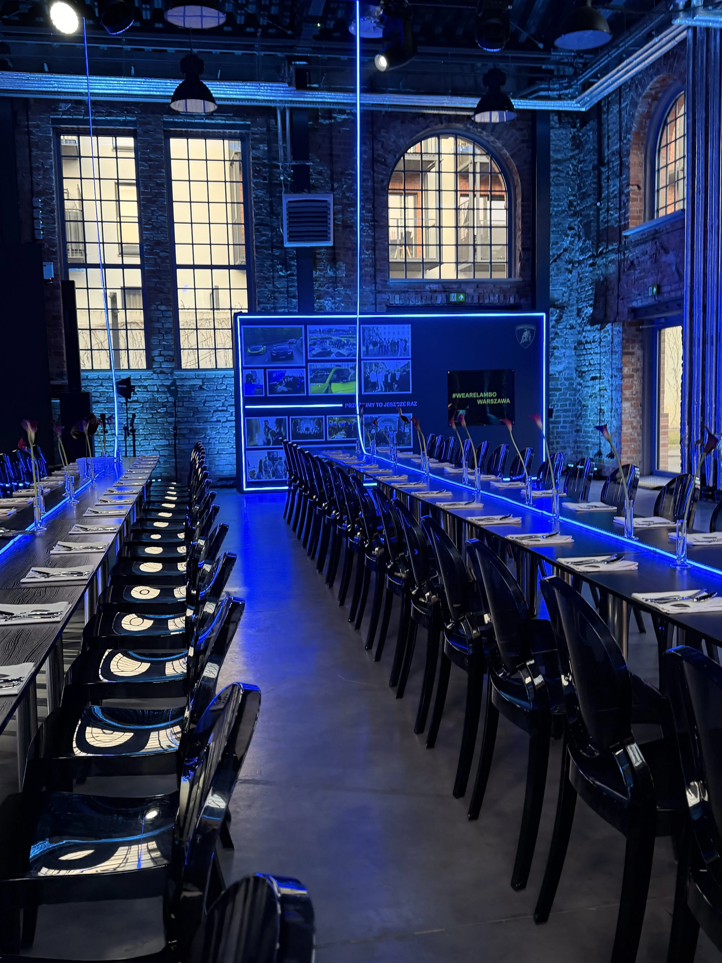 A modern industrial-style dining room with long tables and black chairs set for a meal. The room has large arched and rectangular windows with black frames and a brick wall interior. Blue neon lighting highlights the ceiling and decor, creating a vib