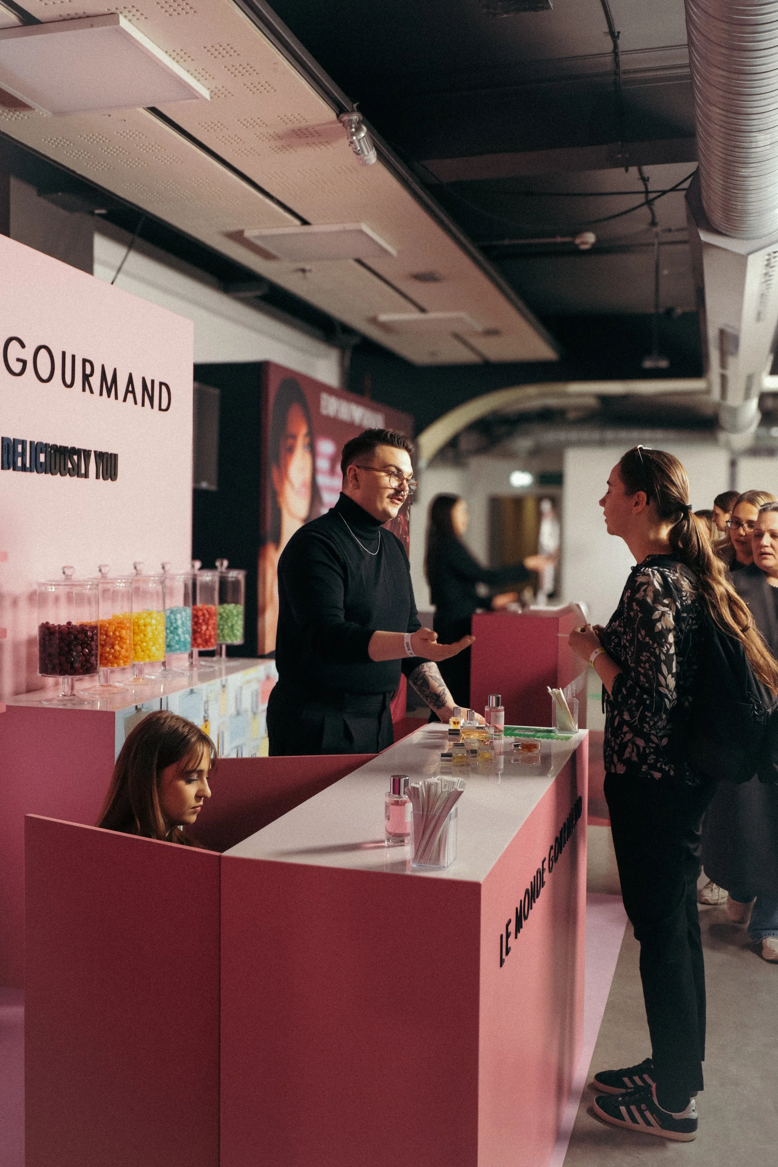 A pink counter at a cosmetics or skincare booth with a woman in a floral top talking to a man in black with glasses, a tattoo, and a necklace, inside a large indoor venue with people in the background.