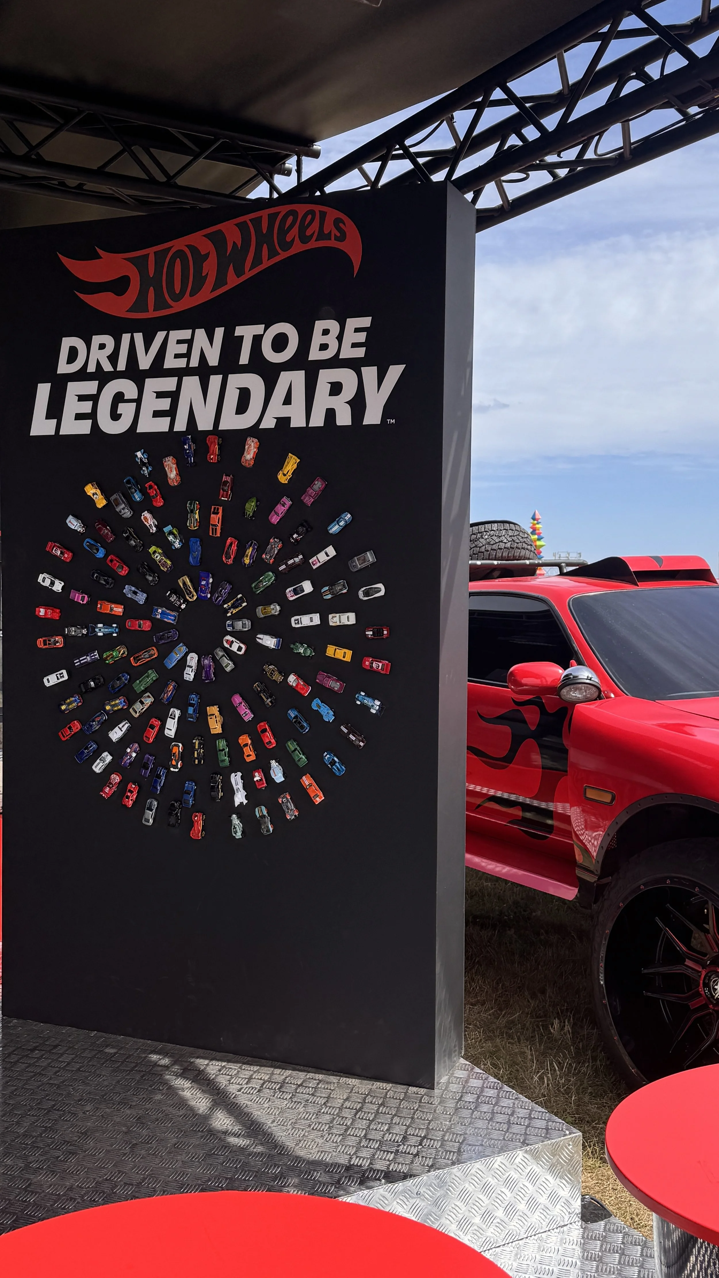 A Hot Wheels display with a sign reading 'Driven to be Legendary' and numerous small toy cars arranged in a spiral pattern on a black background. A red Hot Wheels car is visible in the background.