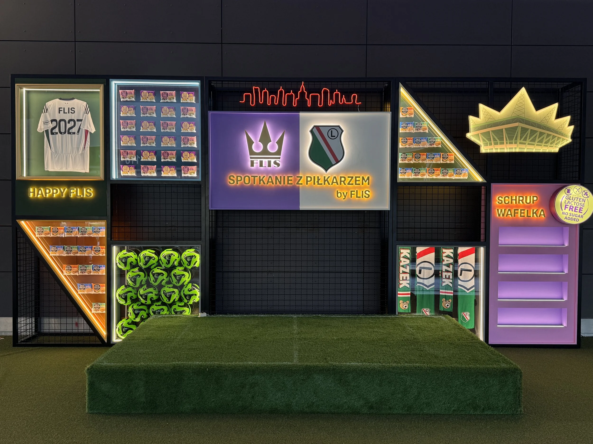 Neon sign of a city skyline, a white football jersey with 'FLIS 2027', shelves of snack packages, illuminated sports helmets, and signs with Polish text promoting food products.