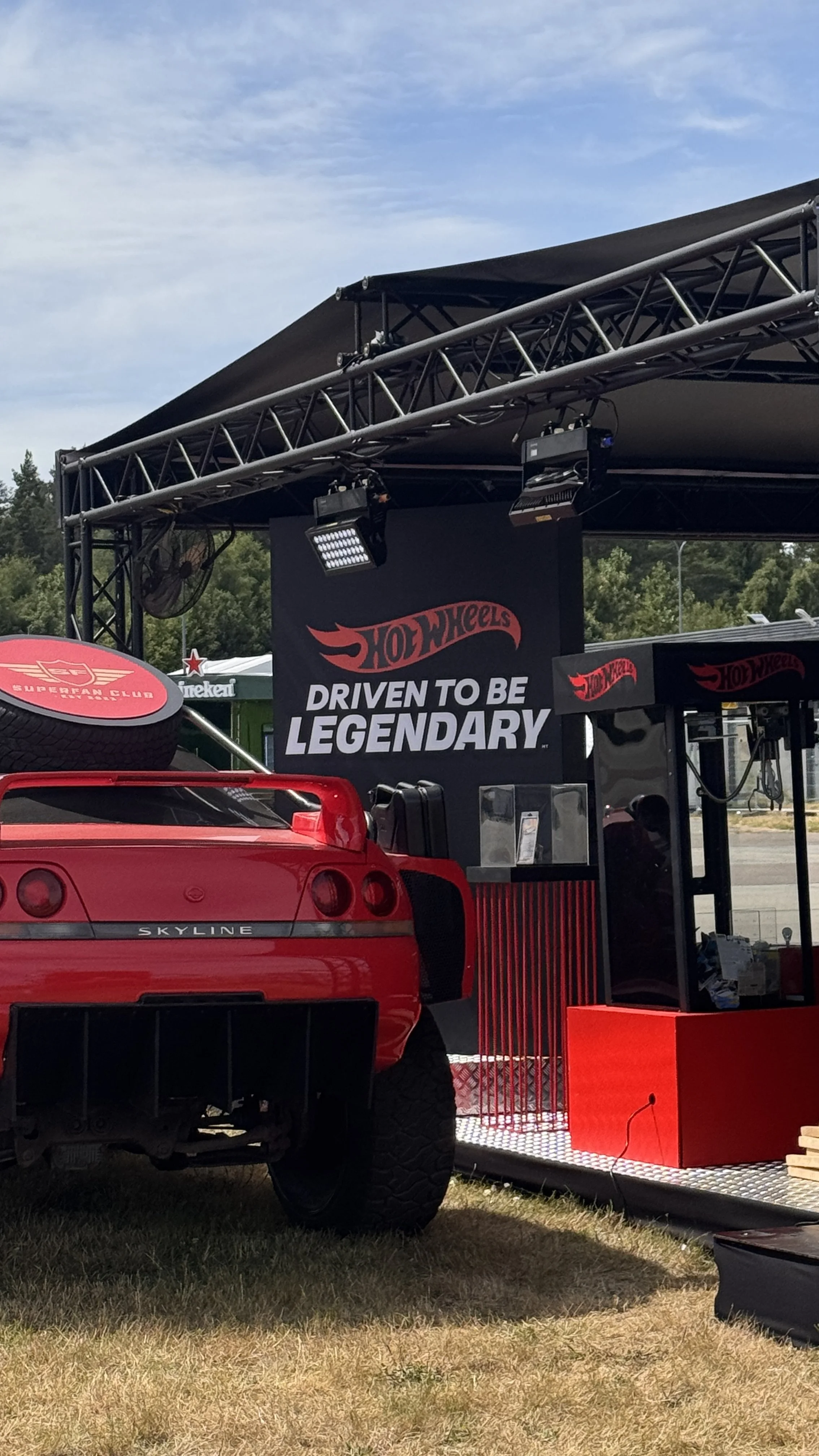 A red Chevrolet Corvette parked in front of a Hot Wheels promotional display with a sign that says 'Driven to be Legendary' and a Hot Wheels logo.