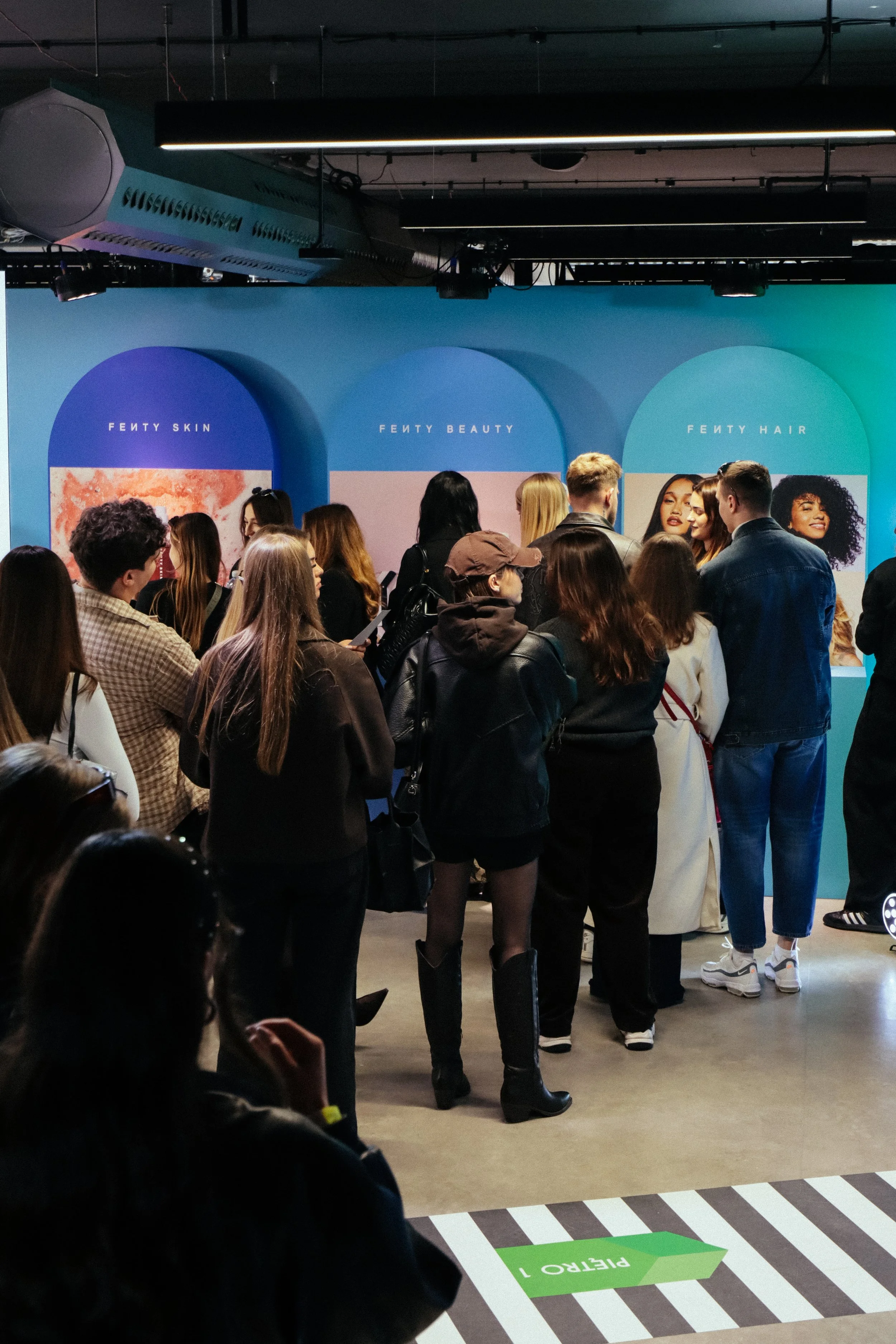 A group of people standing in line at an indoor event with a blue backdrop featuring three arches labeled 'Fenty Skin,' 'Fenty Beauty,' and 'Fenty Hair.'