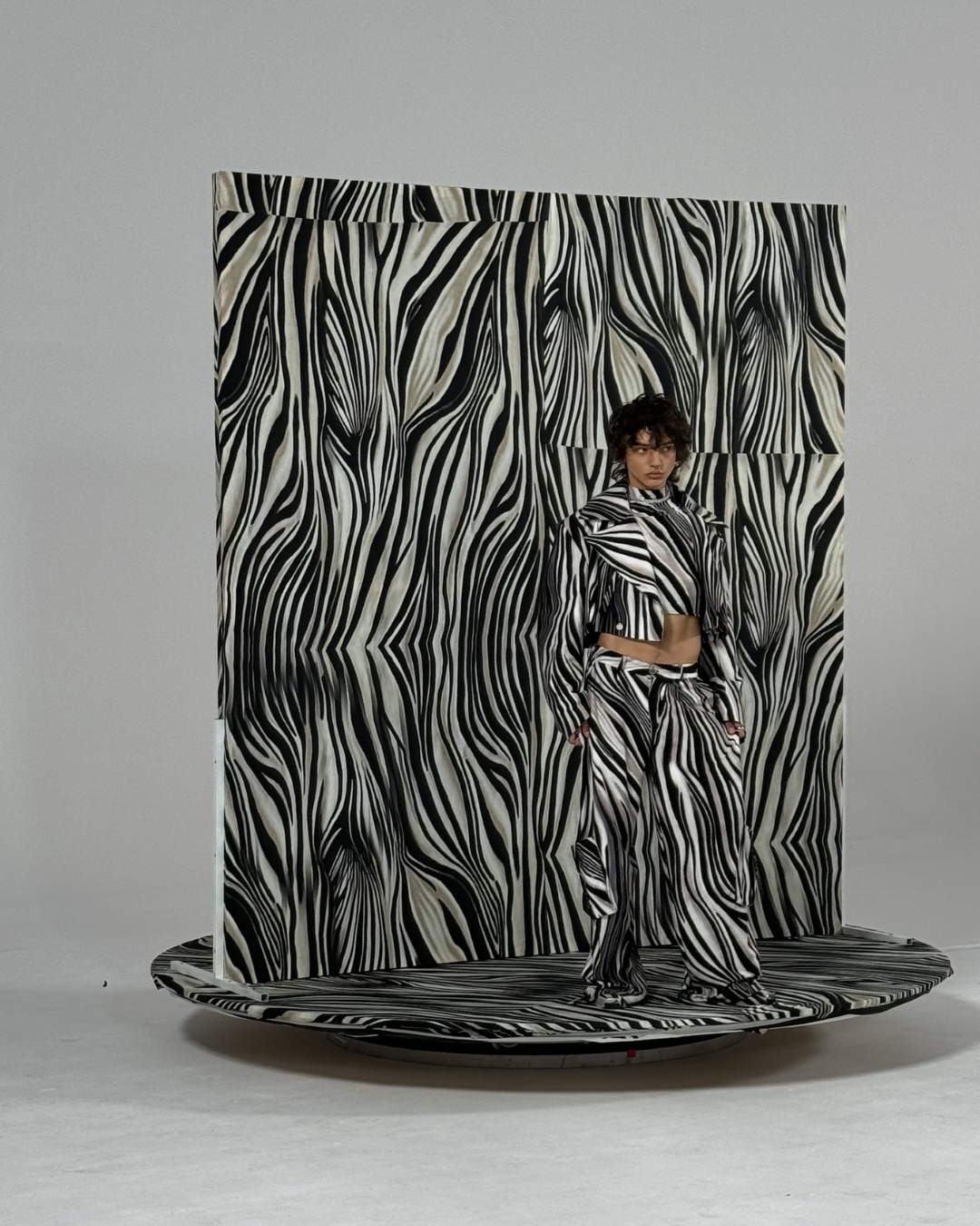 A woman standing in front of a zebra-striped backdrop and platform, wearing a matching zebra-patterned outfit with a cropped top, jacket, and loose pants.