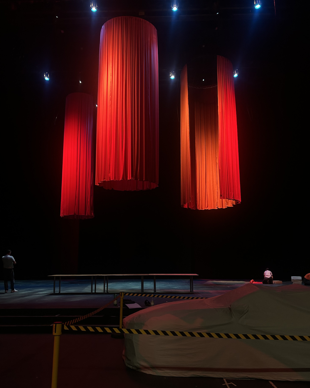 Stage with three large red curtains hanging from the ceiling, lit by blue spotlights, with two people sitting on the floor and one person standing near the stage in a dimly lit auditorium.