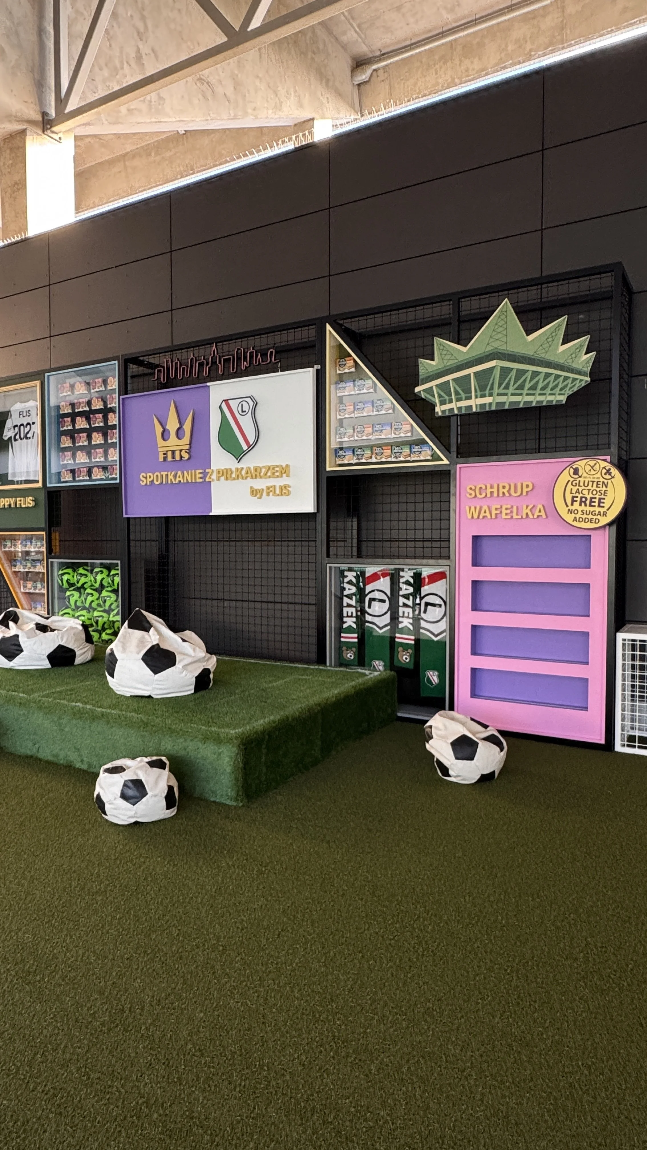 Indoor area decorated with soccer-themed items, including bean bags shaped like soccer balls, a green turf platform, and panels with references to football teams, along with signs in Polish and an illustration of a stadium.