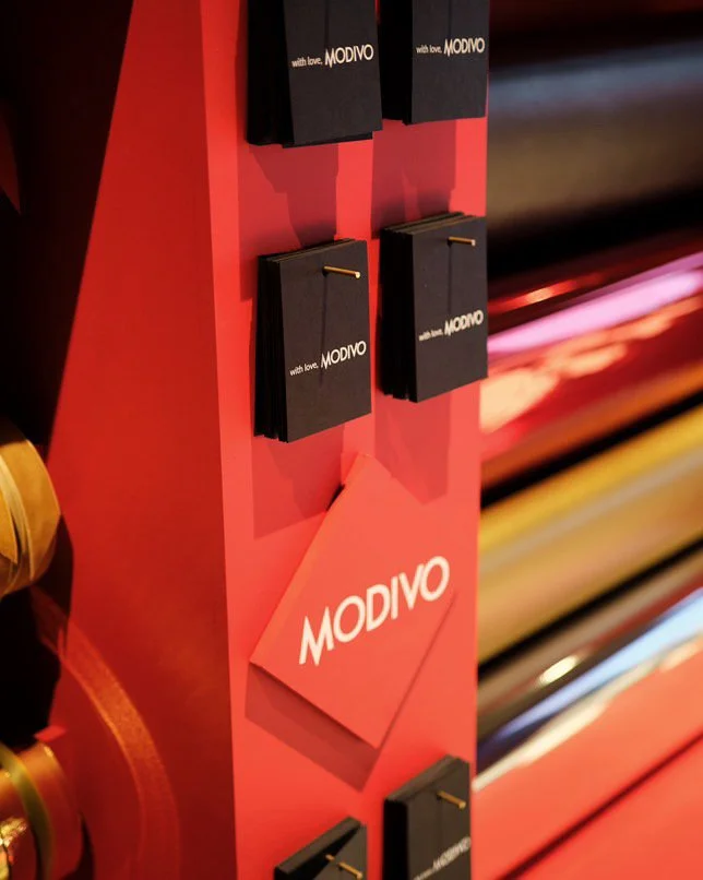 Display stand with black notebooks or pamphlets labeled 'with love, MODIVO' on a red background with the MODIVO logo.