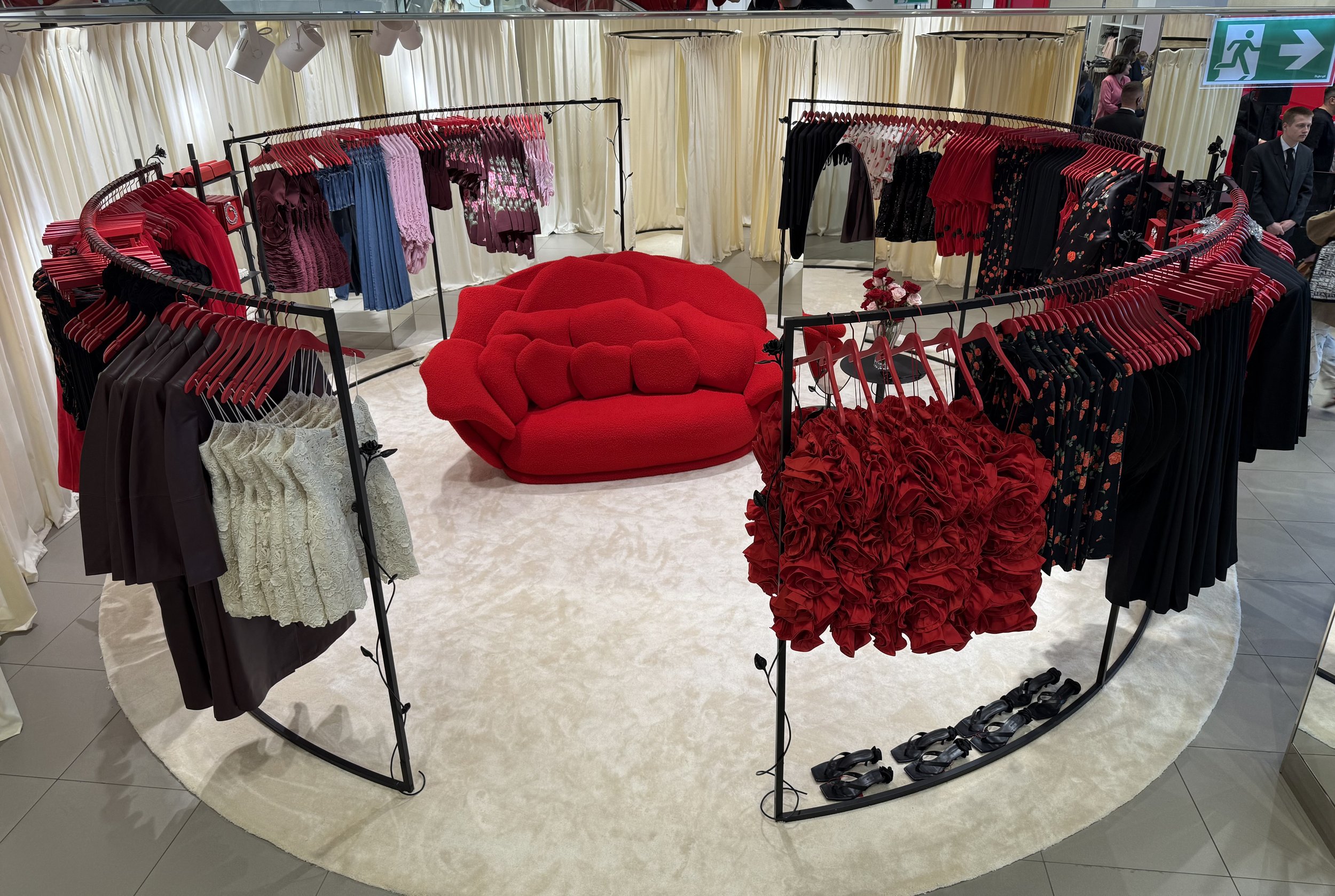 Clothing store display with red, pink, purple, blue, black, and floral dresses on black circular racks. In the center, there is a red sofa and a small round table with pink and red roses. The background has cream-colored curtains and store lighting. 