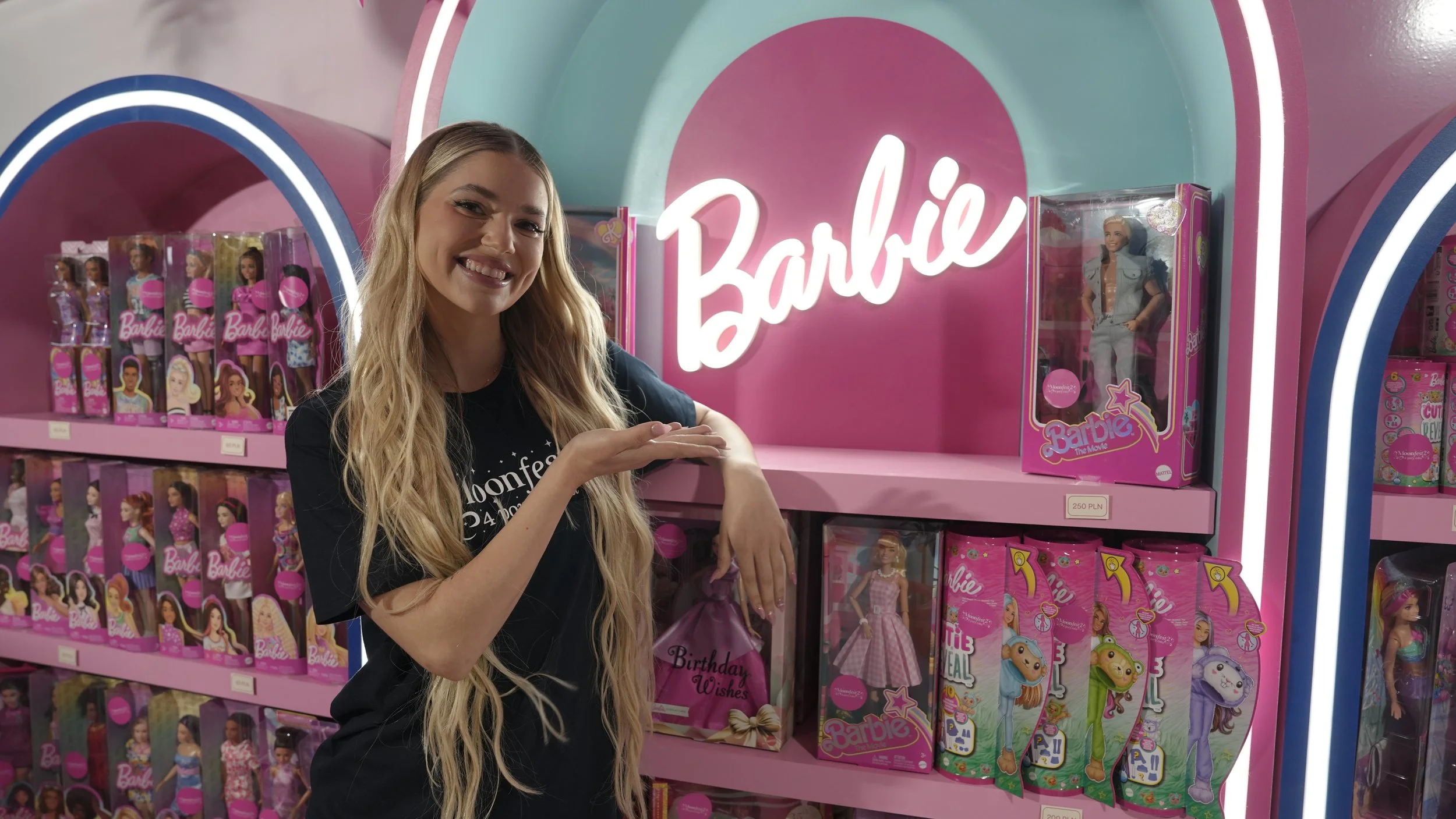 A woman with long blonde hair smiling and showing teeth, standing in front of shelves filled with Barbie dolls and toys at a store or display, with a pink and blue background and a neon-colored Barbie sign.