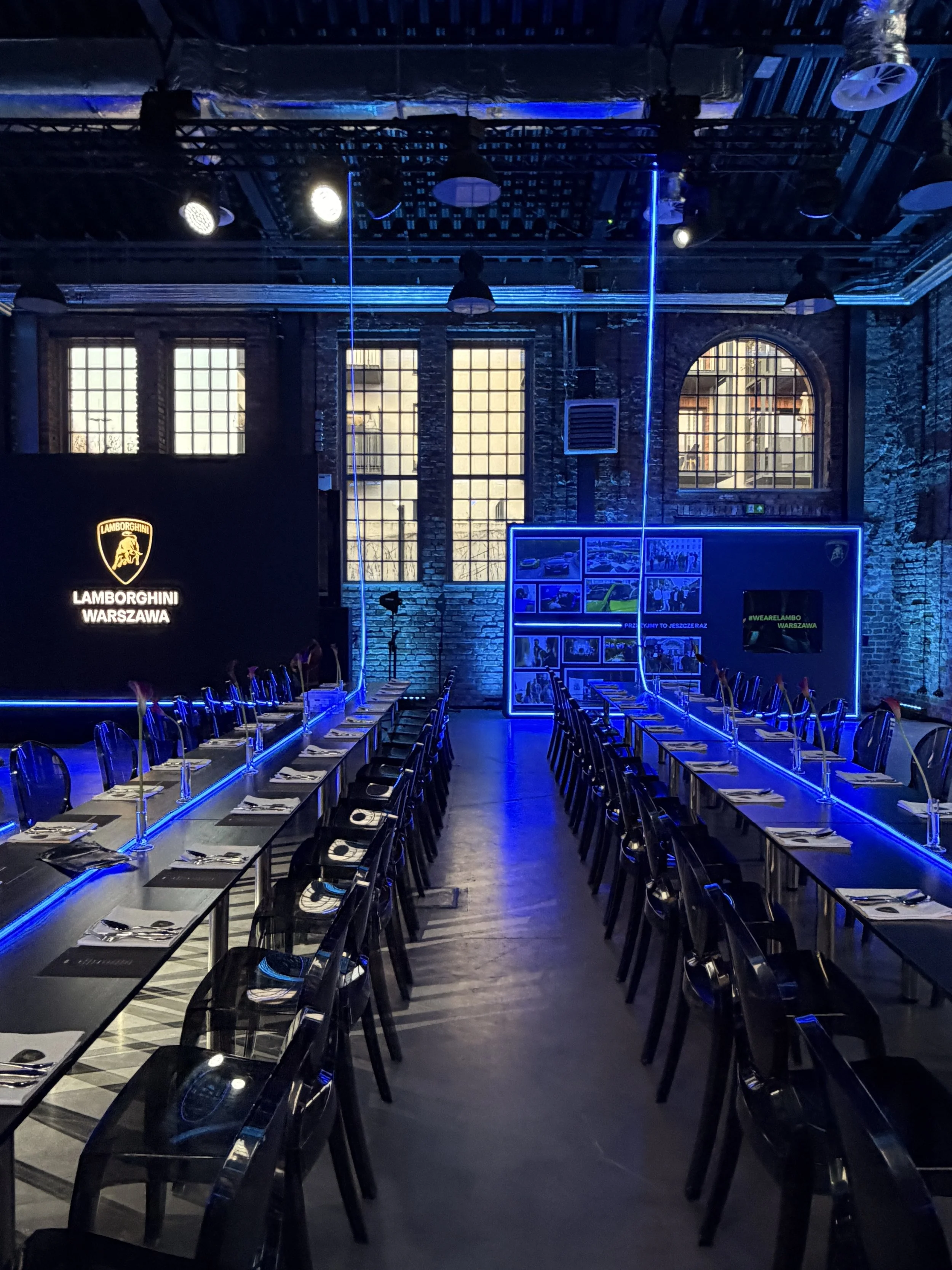 An indoor event space with long tables set for a meal, with black chairs, table settings, and neon blue lighting along the edges. The background features large windows and a brick wall. There is a Lamborghini logo and the text 'Lamborghini Warszawa' 
