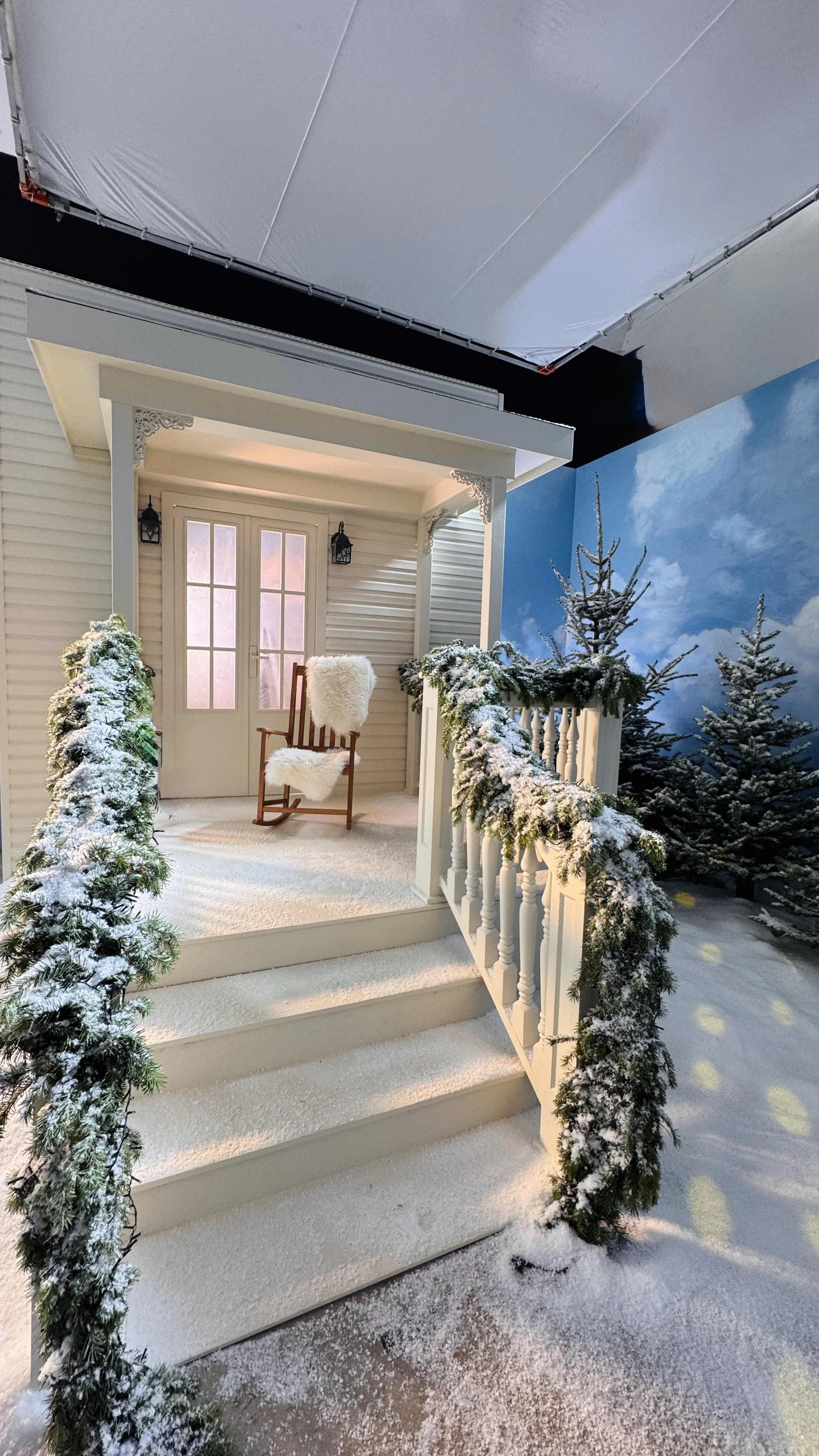 Snow-covered front porch with white steps, decorative green garland, and a small tree. A wooden rocking chair with a furry white cushion is on the porch, and snow is on the ground and trees nearby. The background includes a wall with a painted sky an