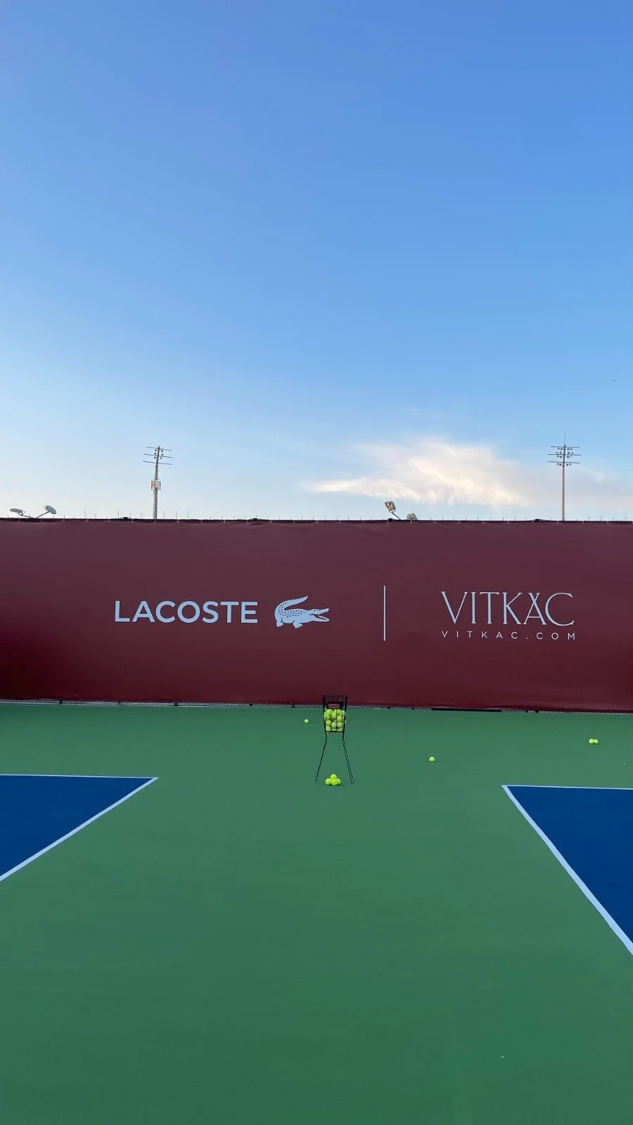 Tennis court with a basket of yellow tennis balls in the center, a few tennis balls scattered around, and a red wall with Lacoste and Vitkac logos. The sky is clear with some clouds.
