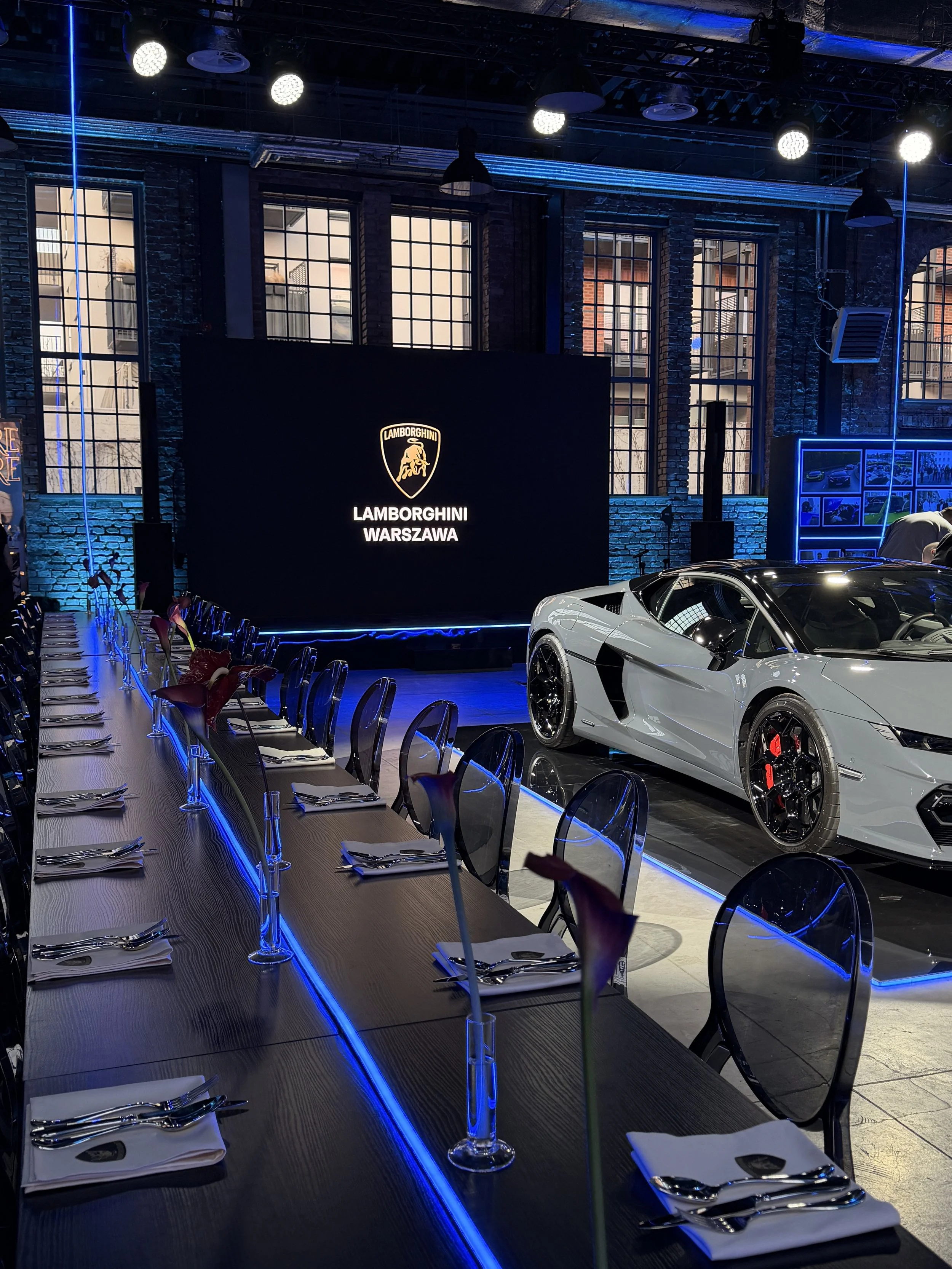A modern event space with a long table set for a dinner, with white napkins and silverware, illuminated by blue accent lighting. In the background, a sports car is on display and a large digital screen featuring the Lamborghini logo and the words 'La