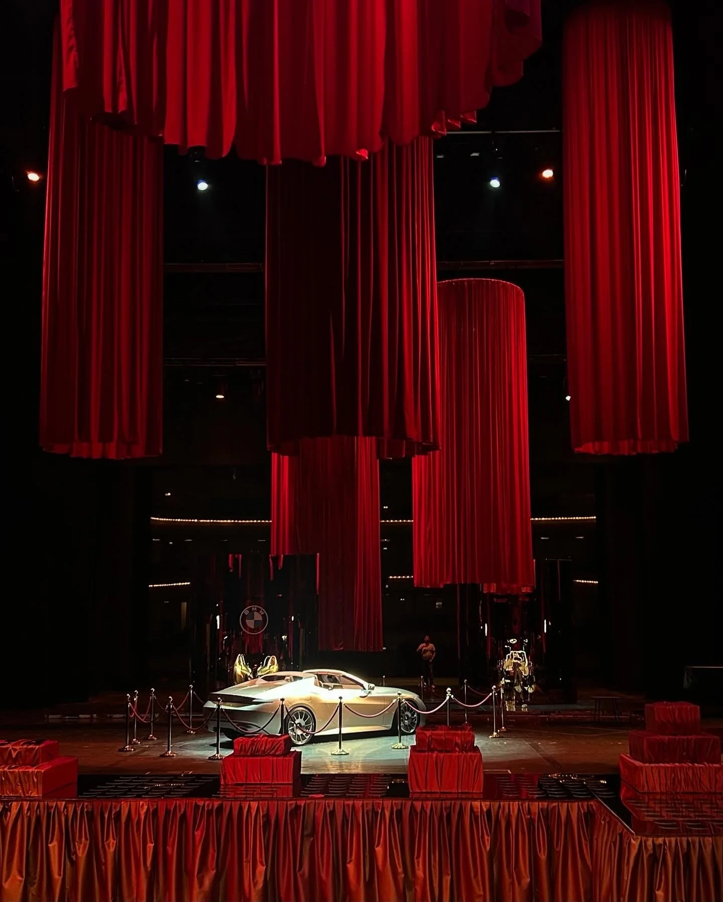 Luxury car displayed on stage with red curtains and velvet ropes around it at an event.