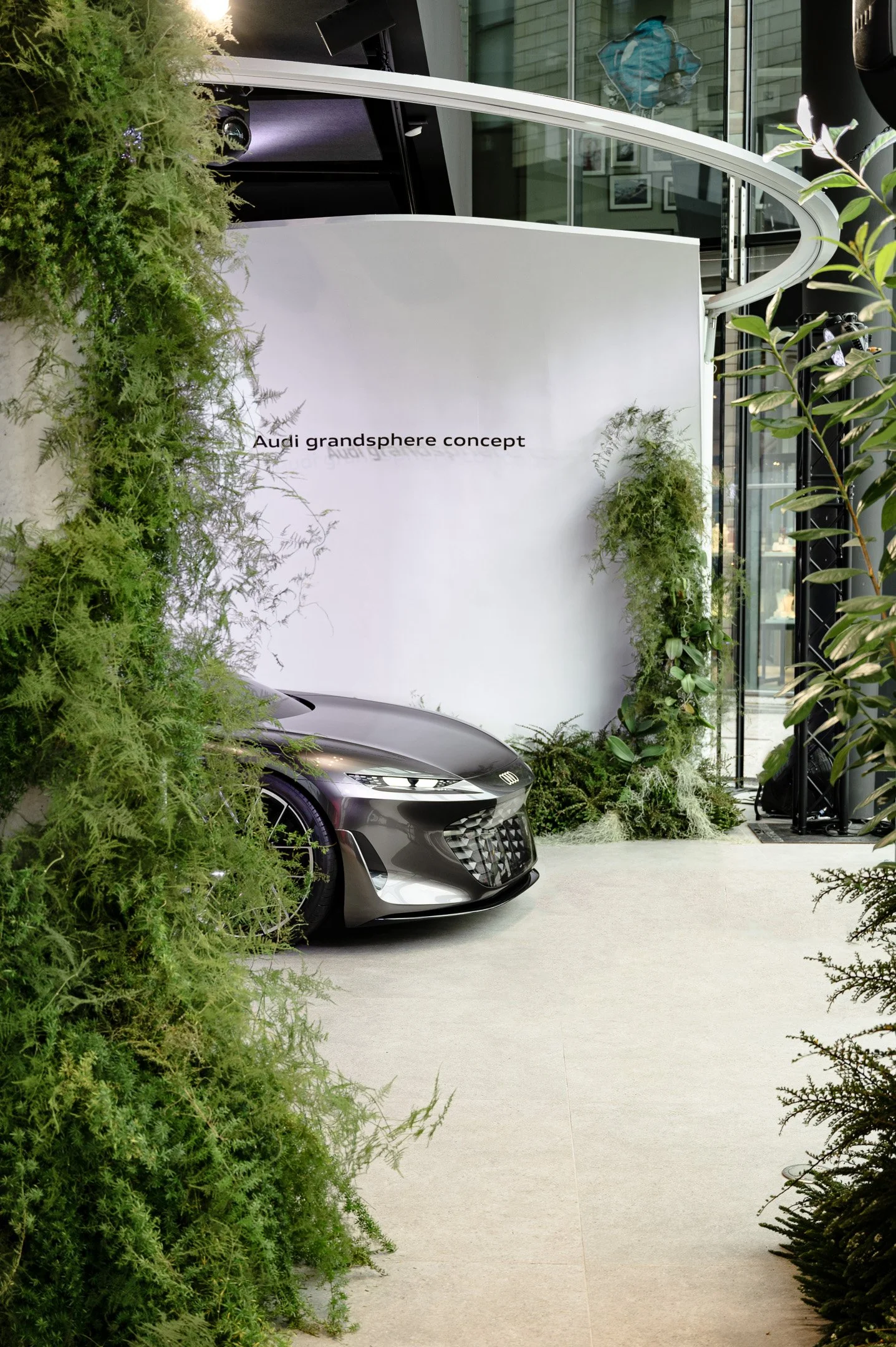 A conceptual display titled 'Audi Gandsphere Concept' features a sleek, dark gray Audi car partially obscured by lush green plants, with a modern building and glass windows in the background.