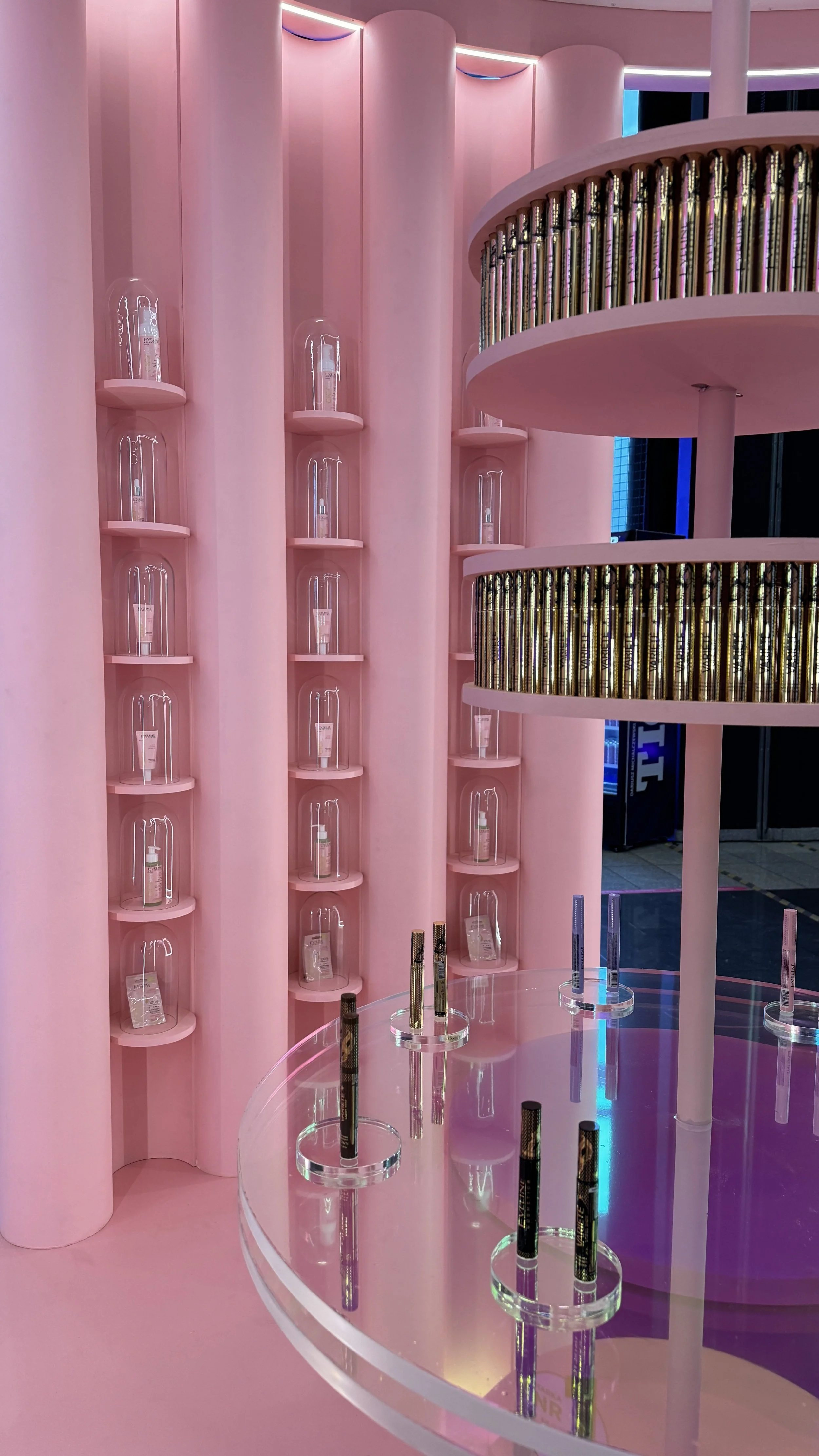Pink interior with cylindrical display shelves holding bottles under glass domes and tubes on a circular glass table, illuminated by colorful lighting.