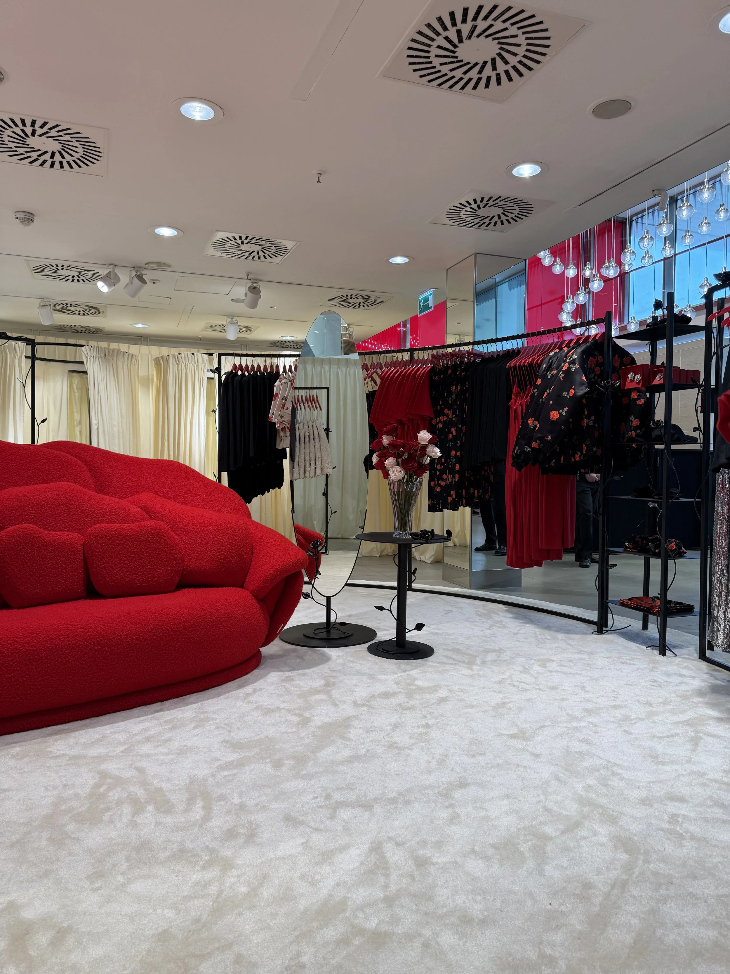 Inside a clothing store with a red sofa, cream curtains, racks of black and red clothing, decorative hanging lights, and a mirror, all on a white carpeted floor.