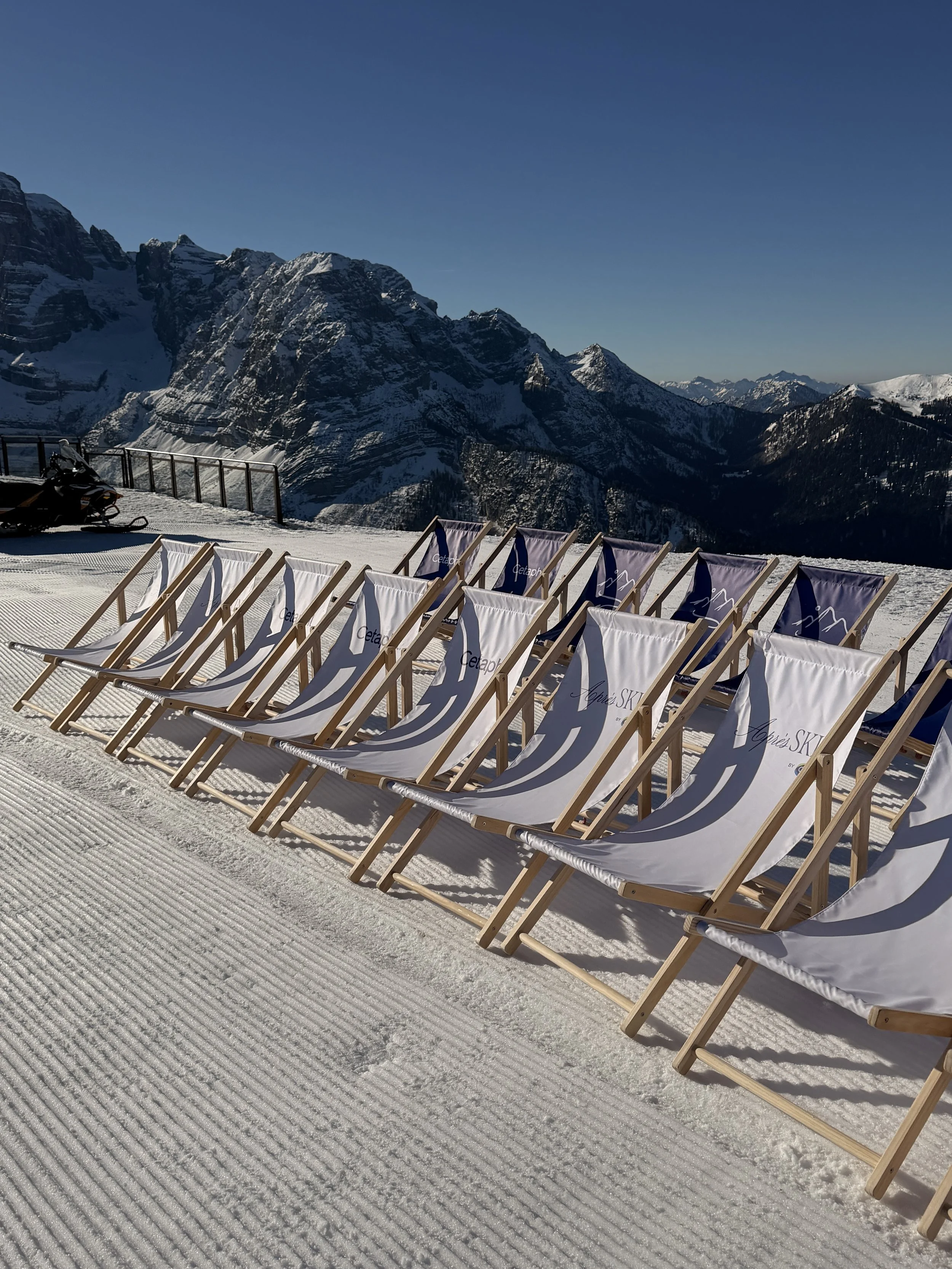 Row of empty lounge chairs on snow-covered ground with mountain range and clear blue sky in the background.
