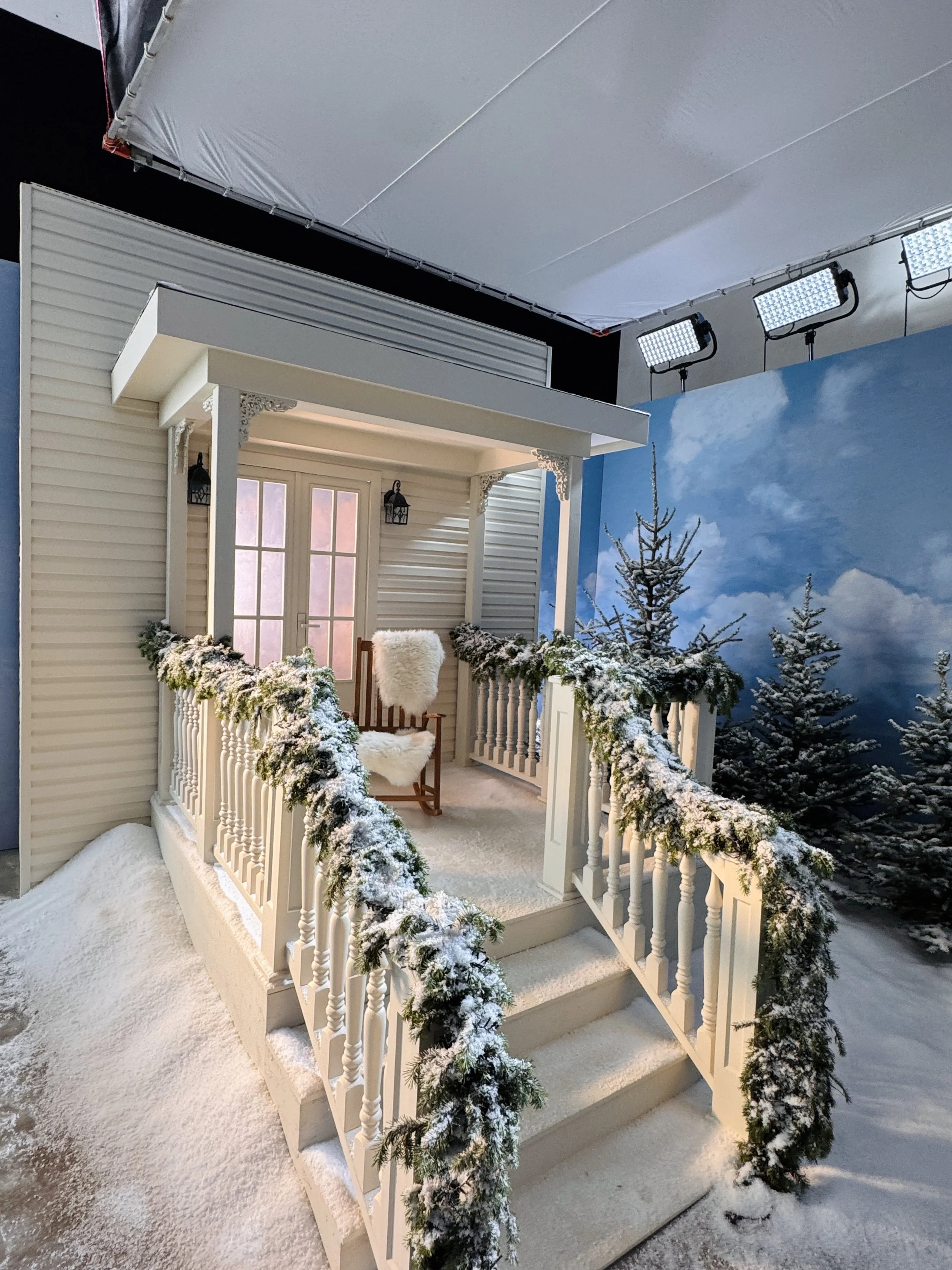Winter-themed porch decorated with snow-dusted green garlands and a white chair, with a backdrop of snowy trees and a cloudy sky mural.