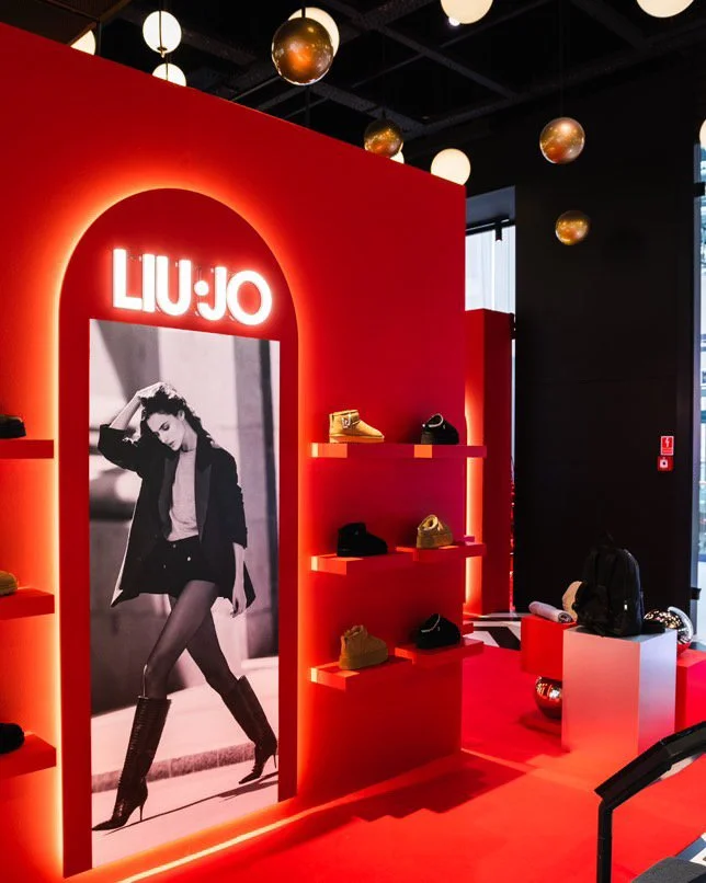Liu Jo retail display with red illuminated wall, black and white fashion photo, and shelves with shoes and handbags, inside a modern store with decorative hanging lights.