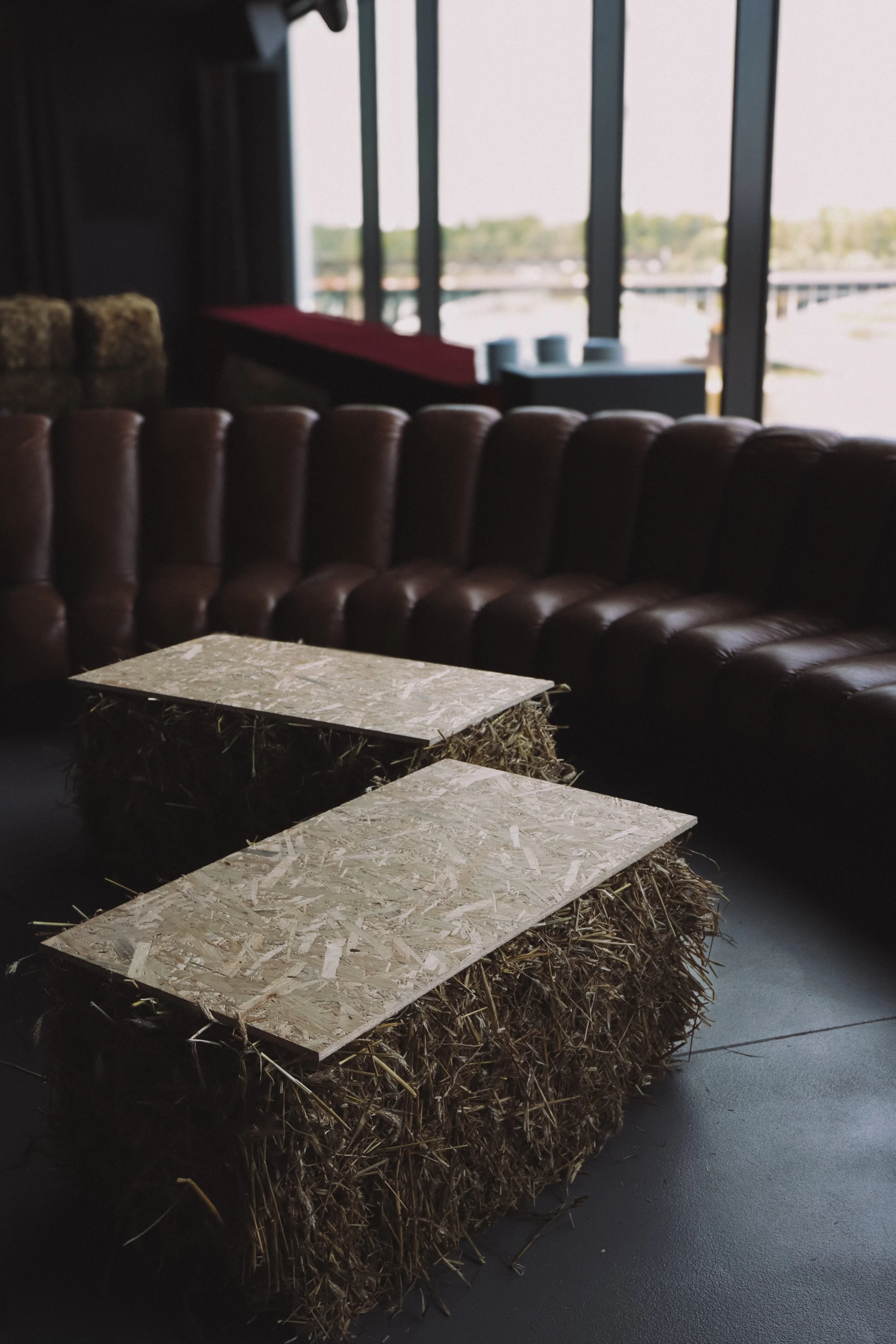 Interior of a modern restaurant or café with a large window view of the outdoors, a brown leather tufted sofa, and a rustic table made of oriented strand board (OSB) panels on a bale of hay.