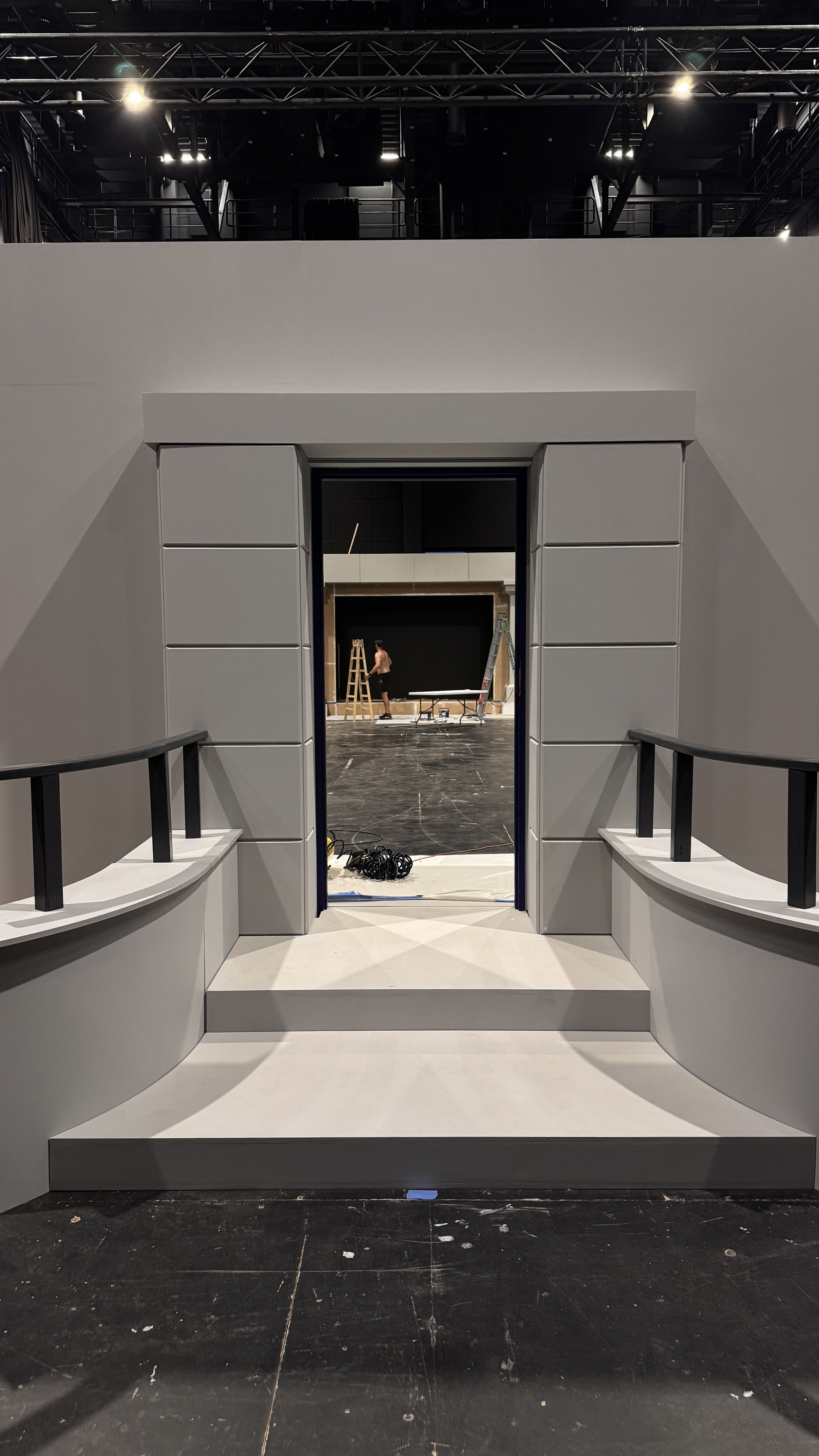View through an opening in a wall leading to a stage or set under construction, with a person working on the stage, ladders, and construction materials visible.