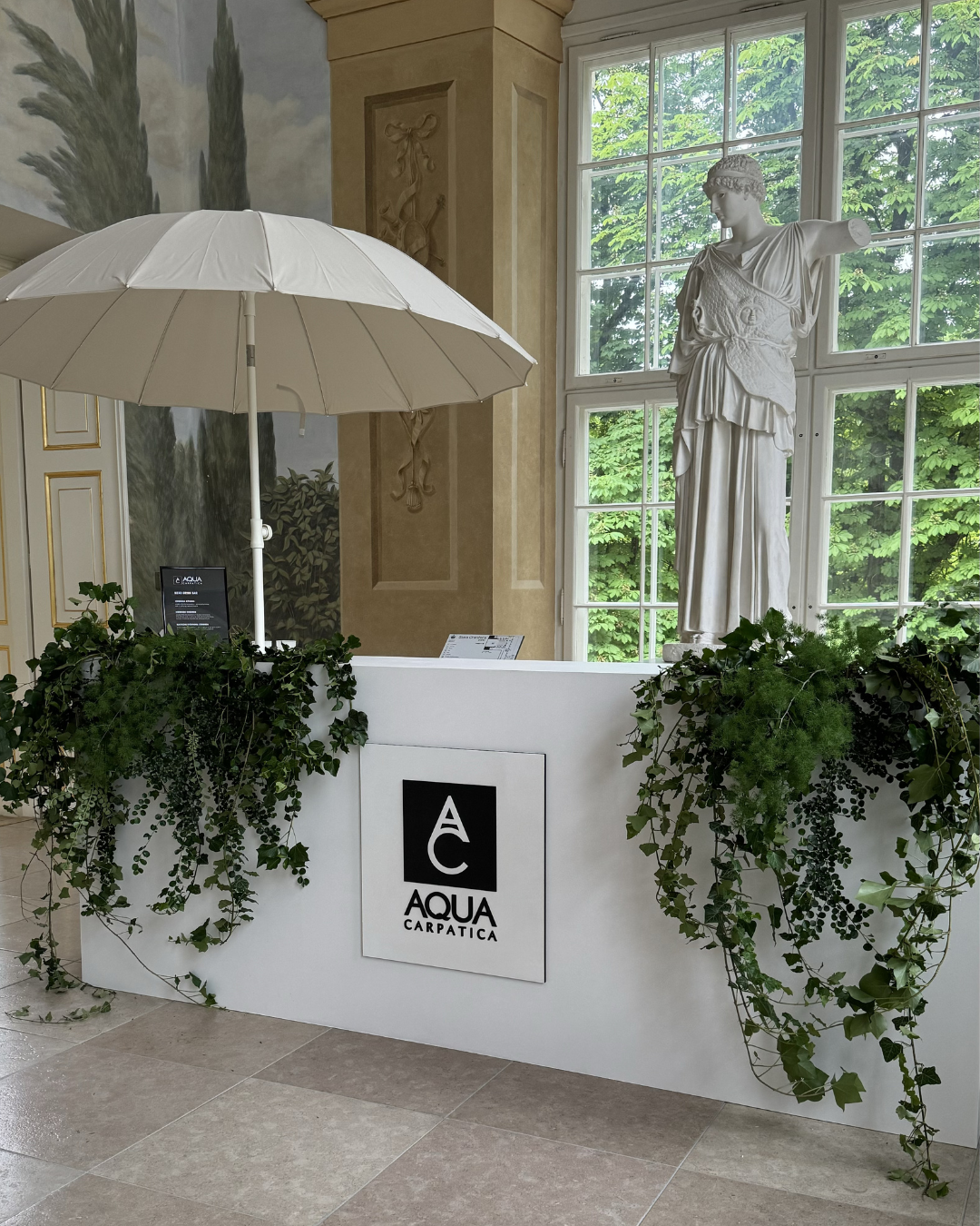 Ancient marble statue of a female figure in classical dress, displayed near a large window with green foliage outside. A white umbrella and a sign with the branding 'Aqua Carpatica' are part of an indoor exhibit with lush green plants.