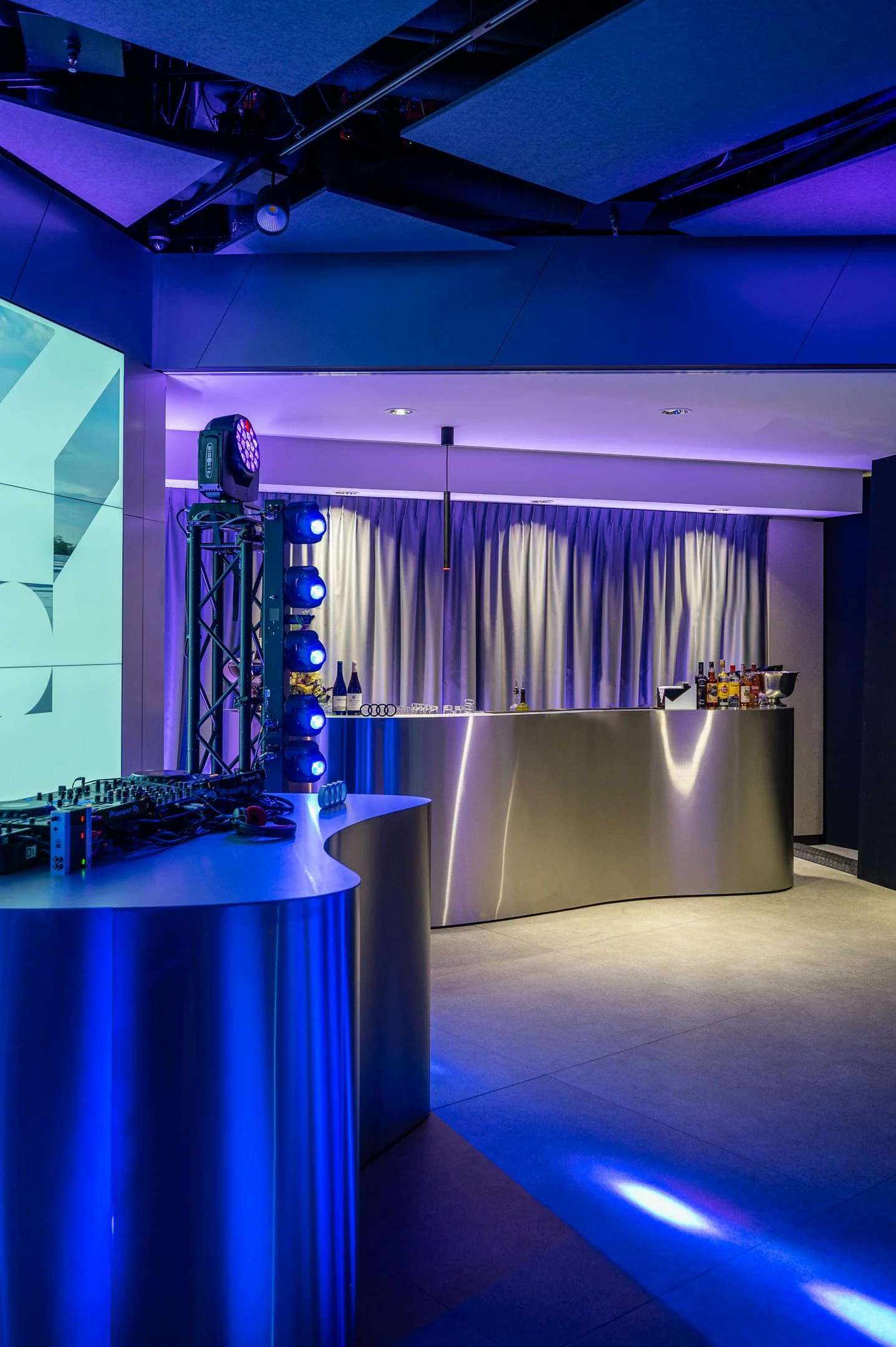 A modern bar with a curved metallic counter, illuminated by blue stage lights. Behind the bar, there are bottles of alcohol and glasses, with purple curtains hanging in the background. The ceiling has dark panels and some stage lighting equipment.