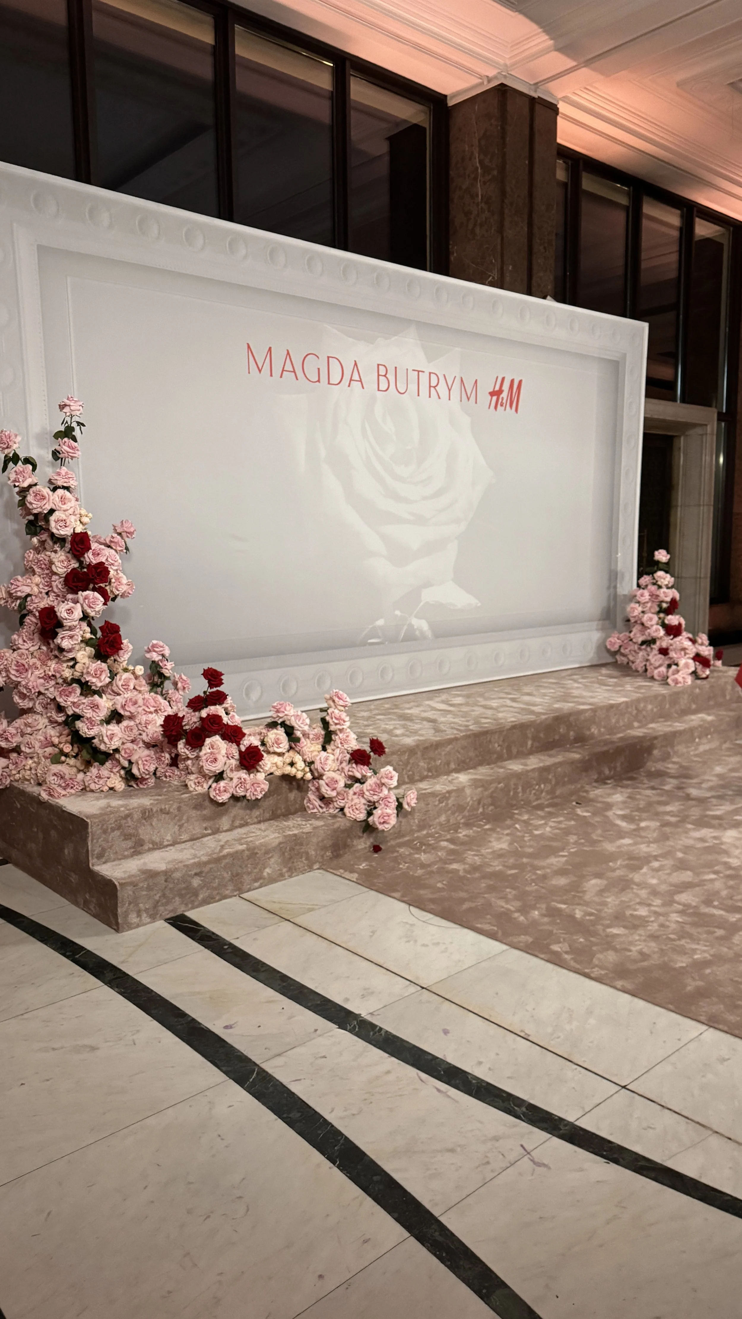 A large white display board with the words 'Magda Butrym H&M' written in red, surrounded by pink and red flowers on a beige carpeted platform in an indoor setting.