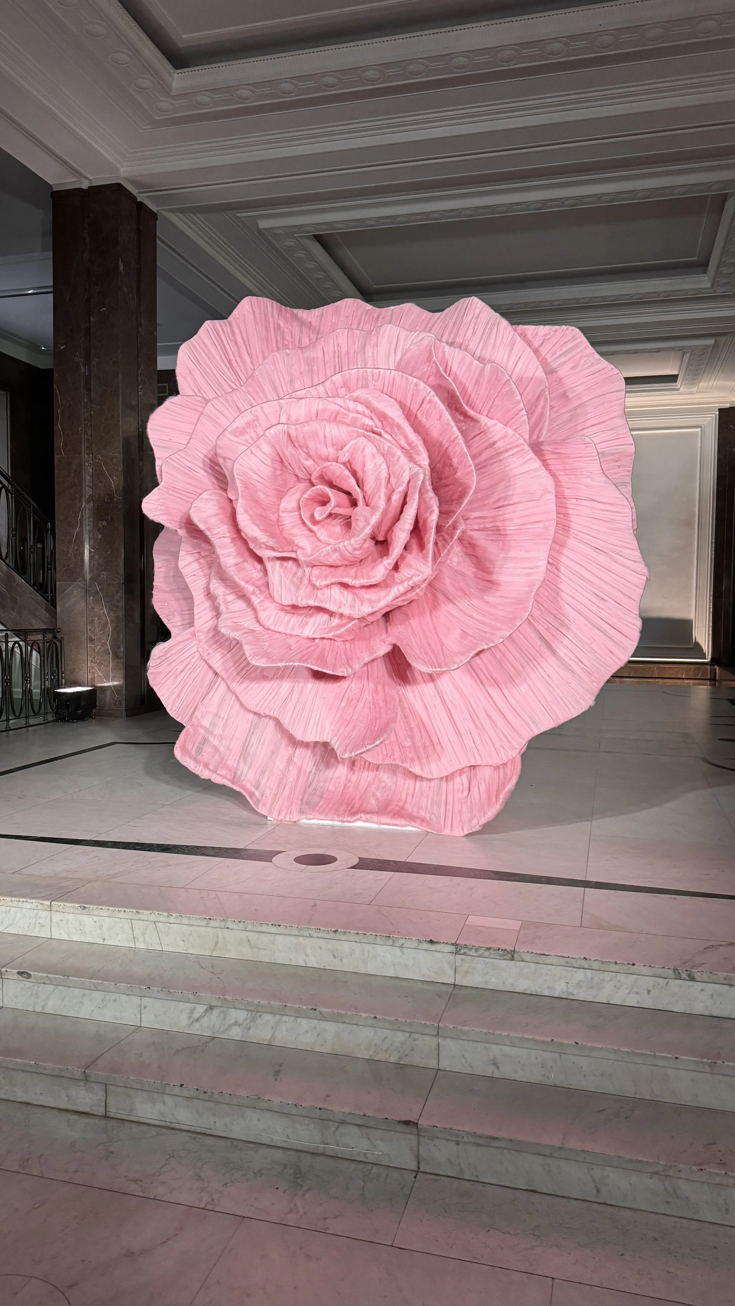 A large pink flower sculpture, resembling a rose, situated inside a building with marble stairs and decorative ceiling moldings.