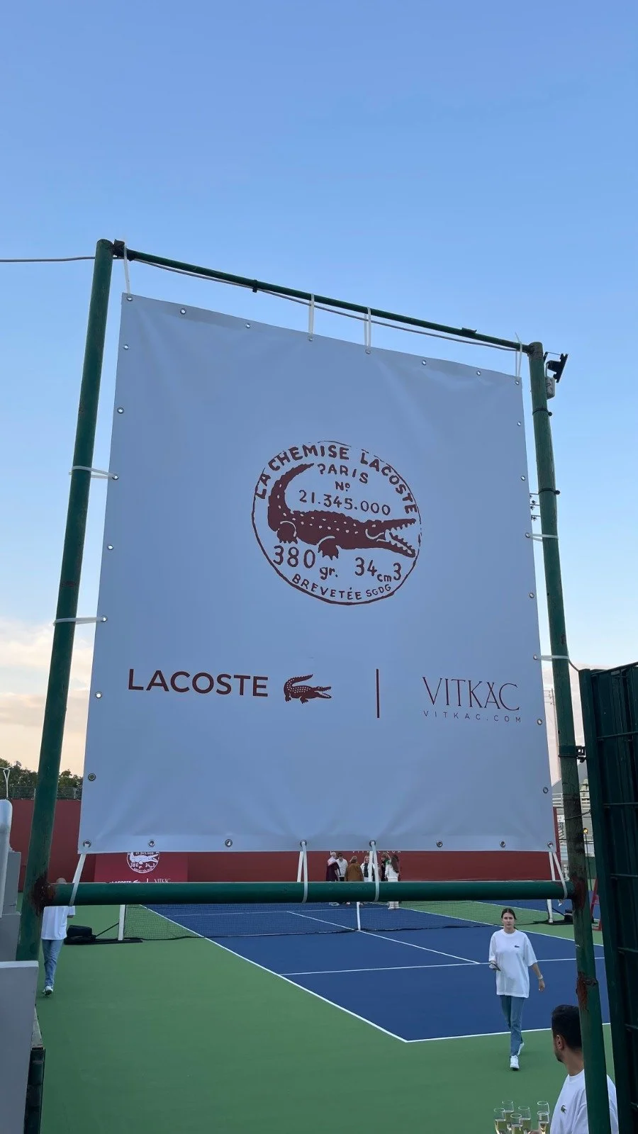Photo of a tennis court with a large Lacoste advertisement banner hanging above it, featuring the iconic crocodile logo.