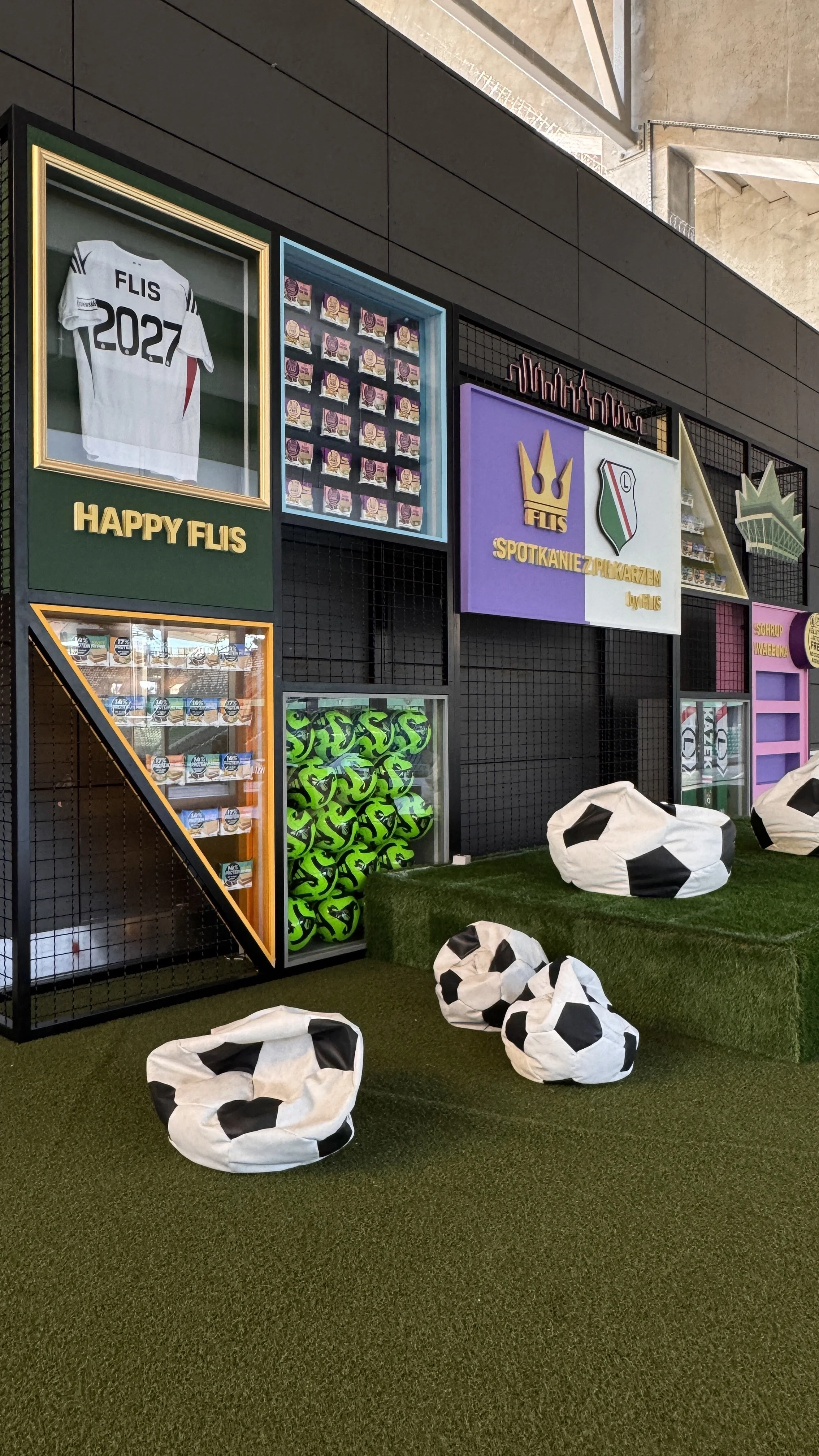 Decorative display of soccer-themed items including a white jersey with 'FLIS 2027', several small boxes, green soccer balls, and bean bag chairs resembling soccer balls, set on artificial grass near a wall with colorful posters and signs.