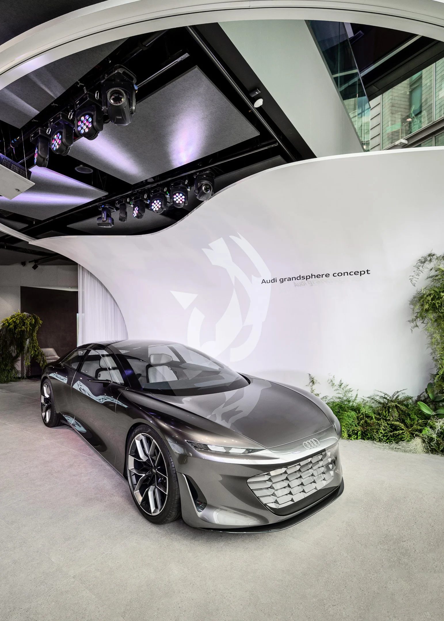 A sleek, silver concept car displayed at the Audi grandsphere concept exhibit, with modern design and futuristic features, parked indoors near a curved white wall with green plants nearby.