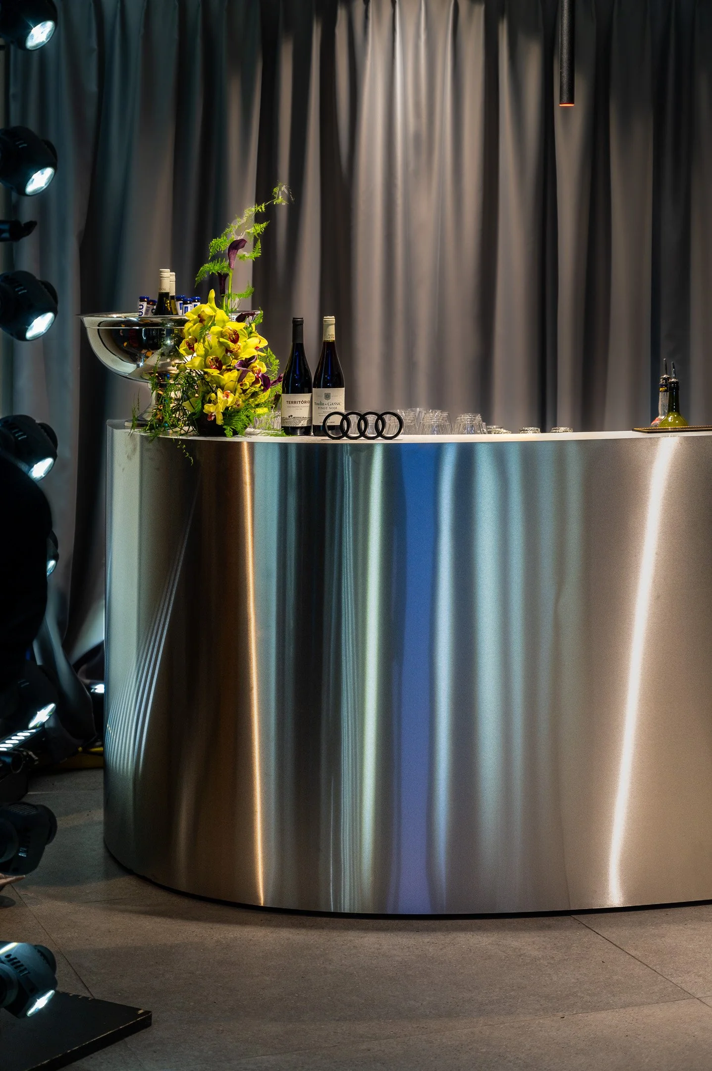 A curved, metallic bar with a flower arrangement, wine bottles, glasses, and an Audi logo, against a dark curtain background with lighting from spotlights.