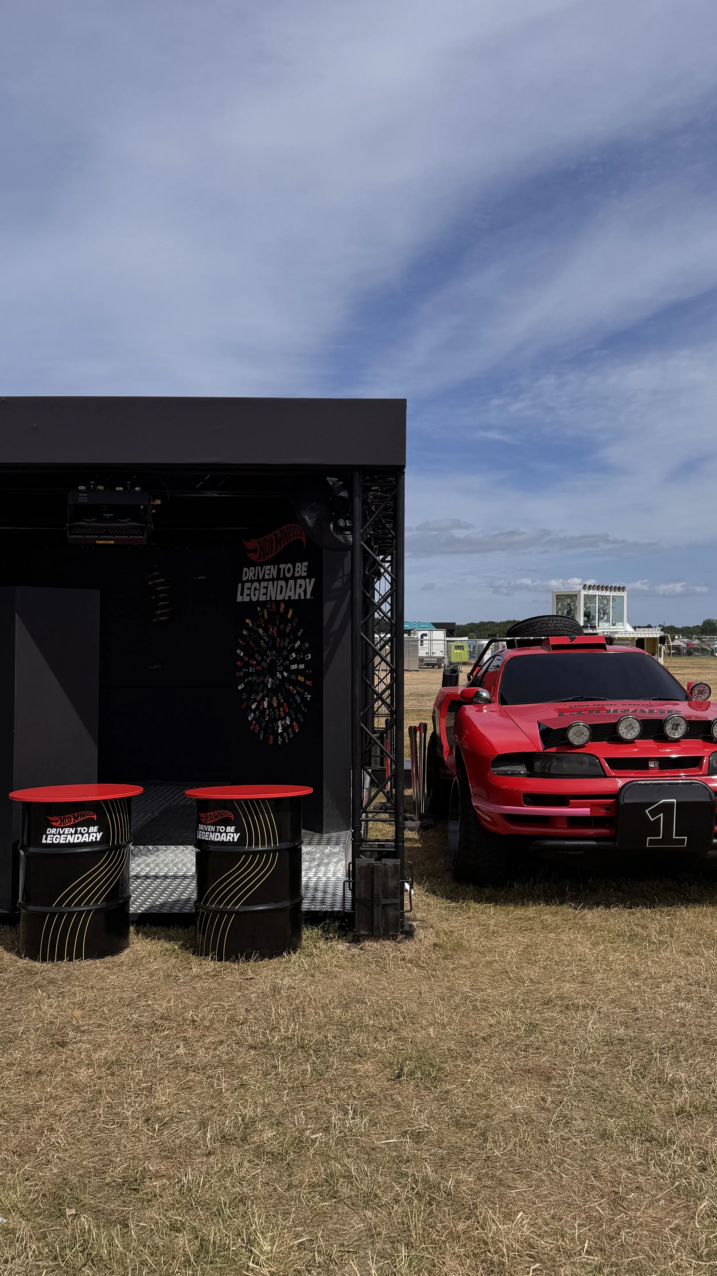A red race car parked next to a hot wheels themed display at an outdoor event. The display includes a black stage with the Hot Wheels logo and the slogan 'Driven to be Legendary', along with two black barrels with similar branding. There are also som