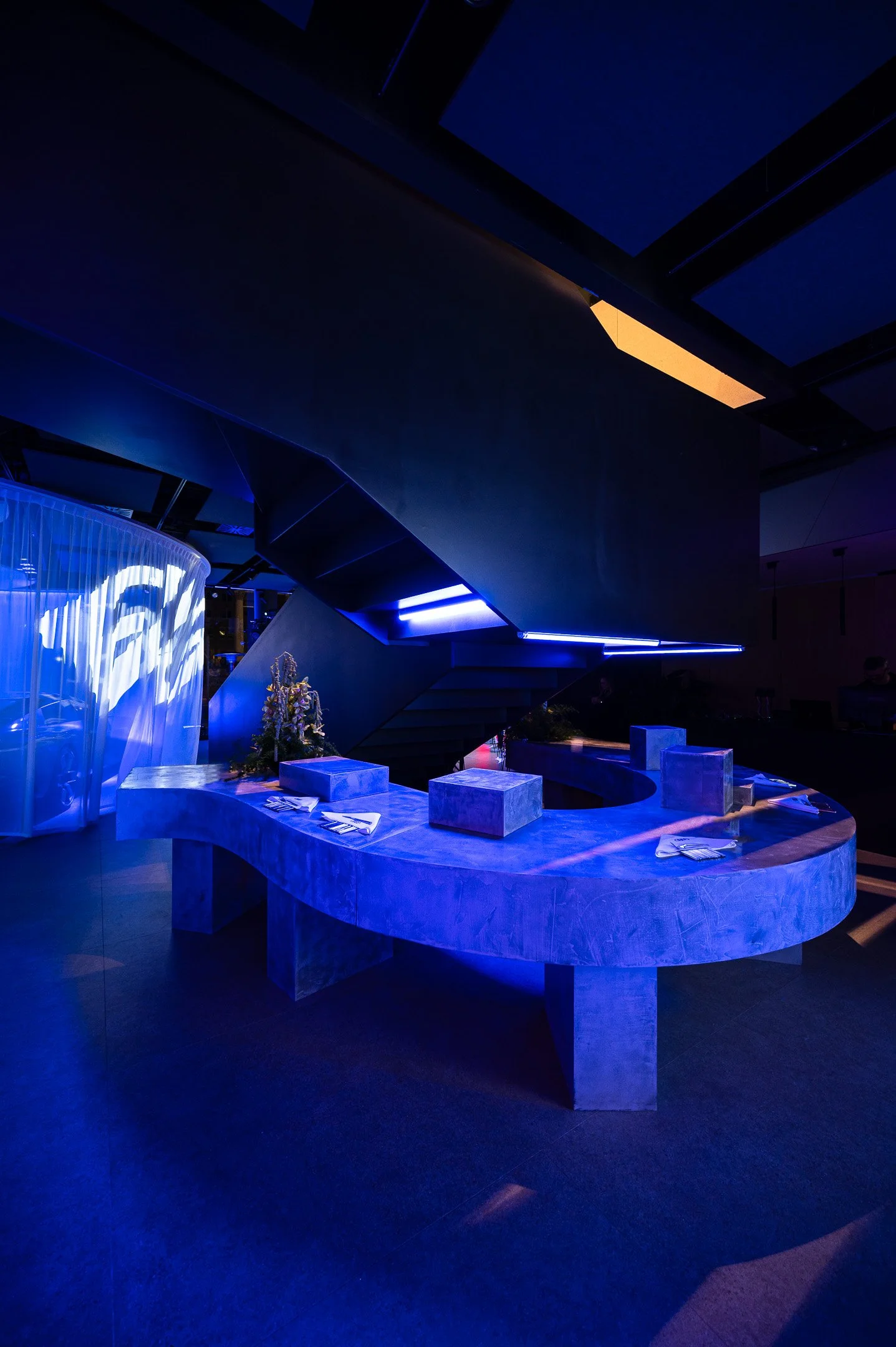 Modern indoor space with a marble curved bar, blue lighting, and a staircase with a blue glow, decorated with flowers and set table.