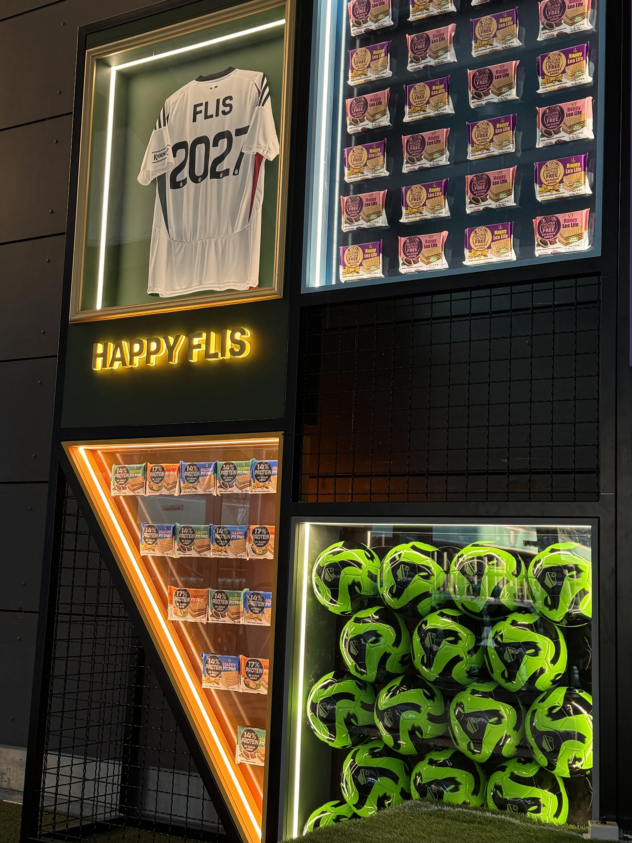 A sports-themed display featuring a framed white soccer jersey with 'FLIS 2024' on the back, illuminated with LED lights. Next to it is a collection of snack bags with colorful packaging and the label 'Happy Lea Life.' The display also includes a clu