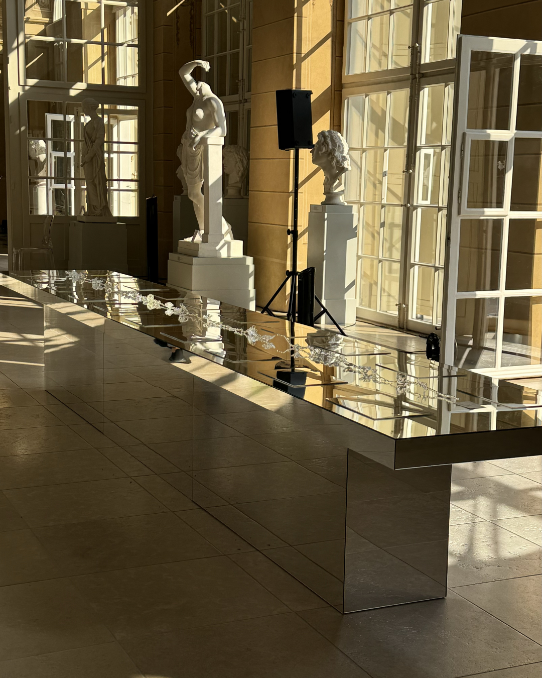 A display of classical sculptures in a bright, sunlit gallery with large windows, a mirrored table with jewelry, and a black speaker on a stand.