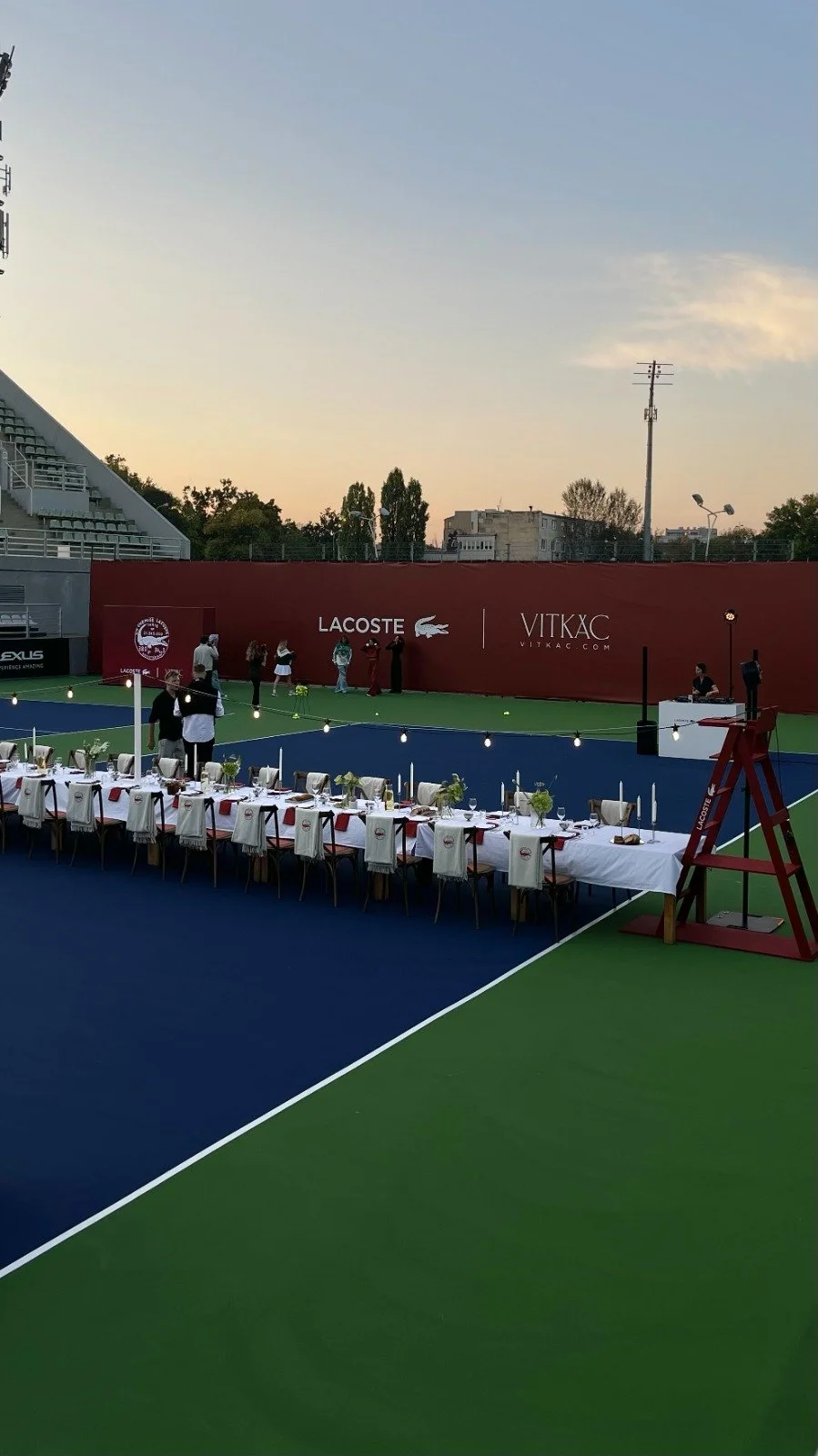 An outdoor tennis court set up for a special event with a long, decorated dining table and people gathering near a red wall featuring Lacoste and Vitkac brands, with the sky fading into sunset.