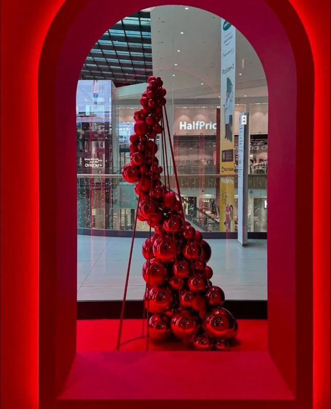 Red Christmas ornaments arranged in a cone shape inside a red archway, displayed in a shopping mall.