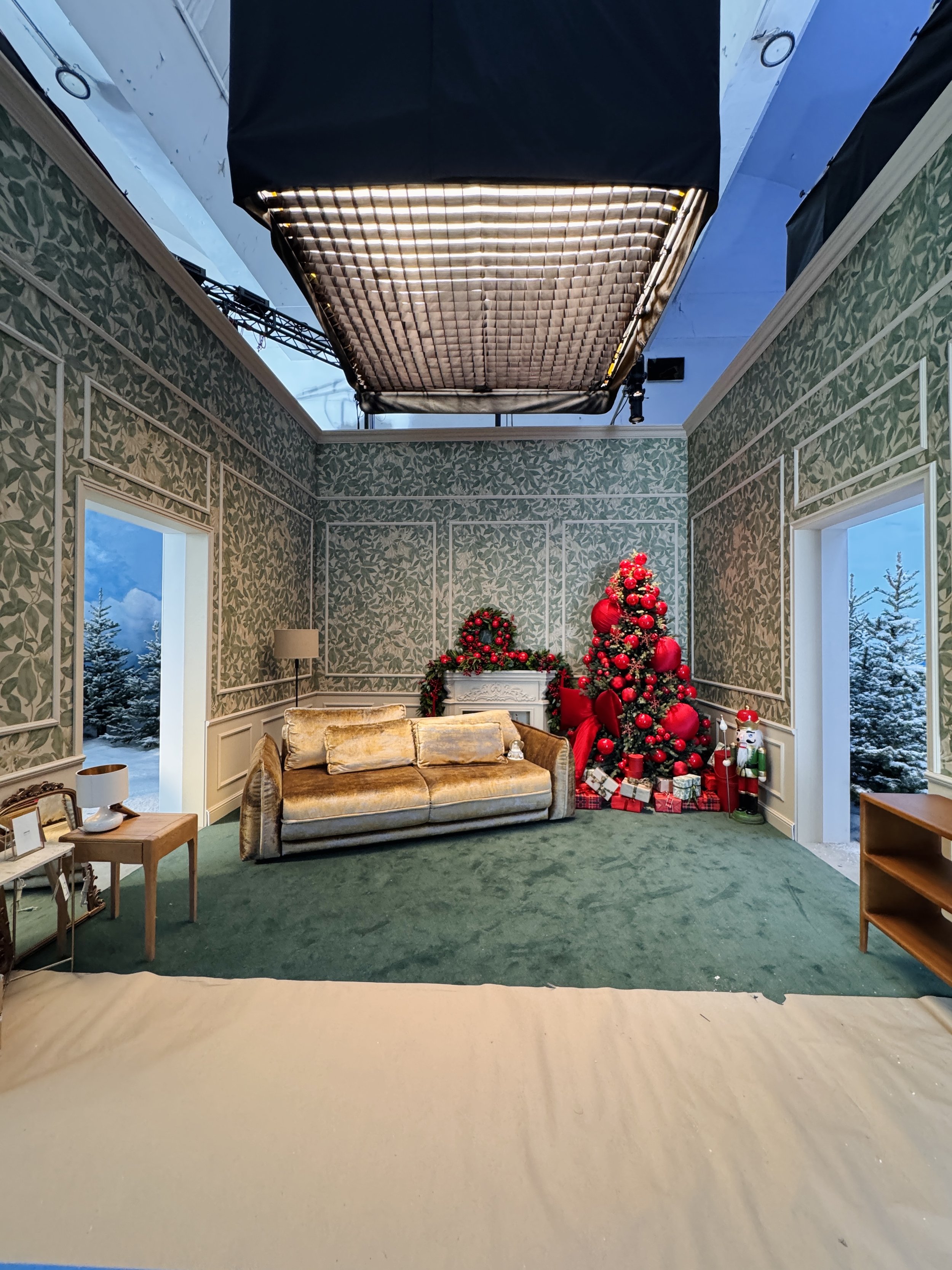 Indoor studio setup with Christmas decorations including a Christmas tree, gifts, and holiday ornaments; contains a backdrop with a snowy outdoor scene viewed through windows; studio lighting equipment overhead; vintage-style sofa in the foreground.