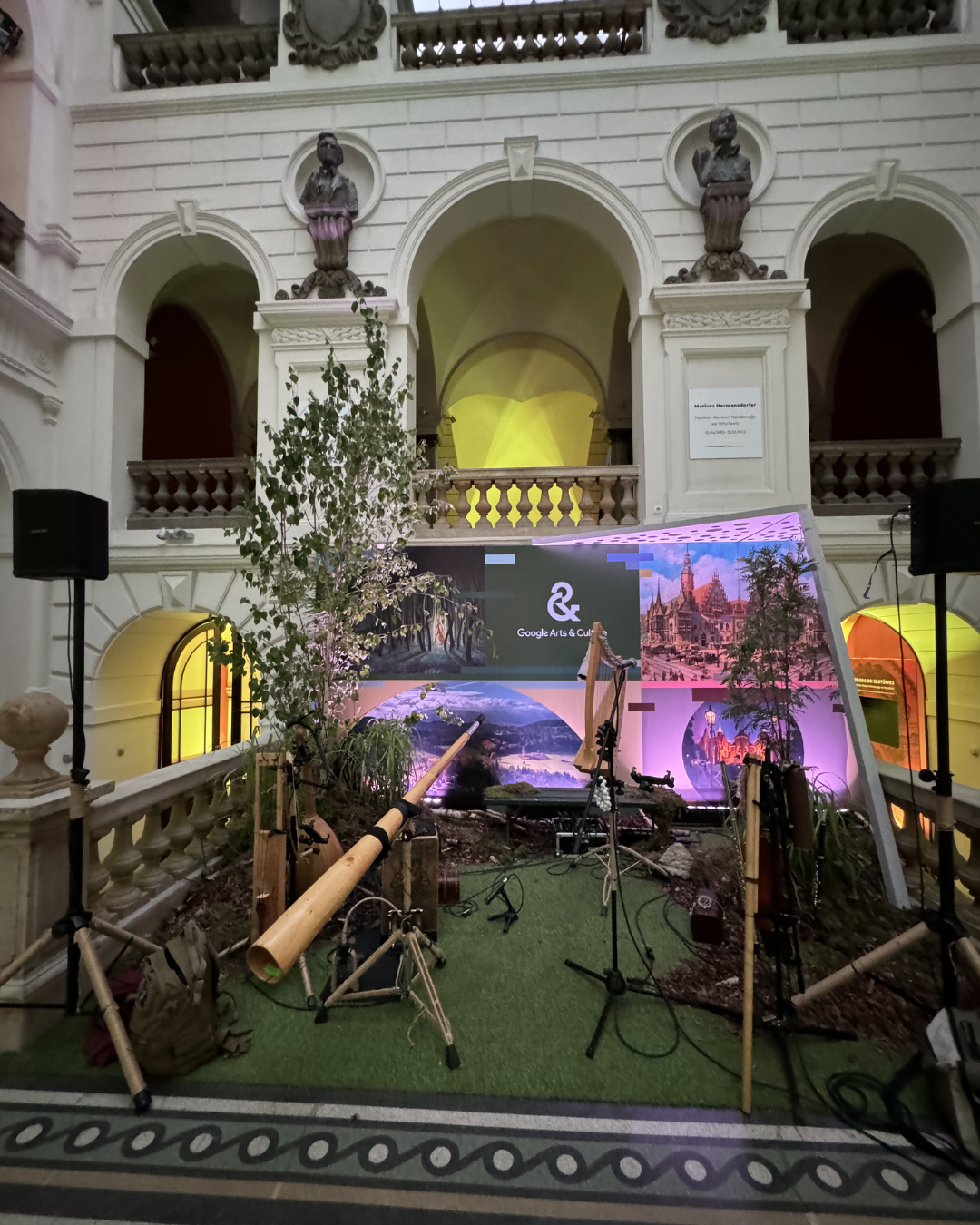 An indoor art installation with musical instruments and microphones on a grassy surface, set against a backdrop of images of landscapes and buildings, inside a grand historic building with classical architecture, statues, and decorative elements.