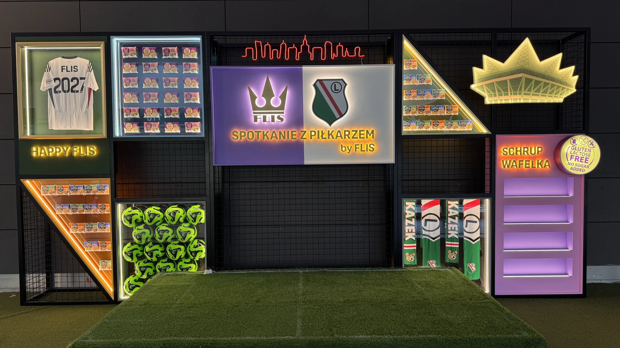 A display with neon signs and sports-themed items, including a white soccer jersey with 'FLIS 2027' on it, various snack packages, neon lights of a city skyline and a crown, green soccer balls, and sports scarves.