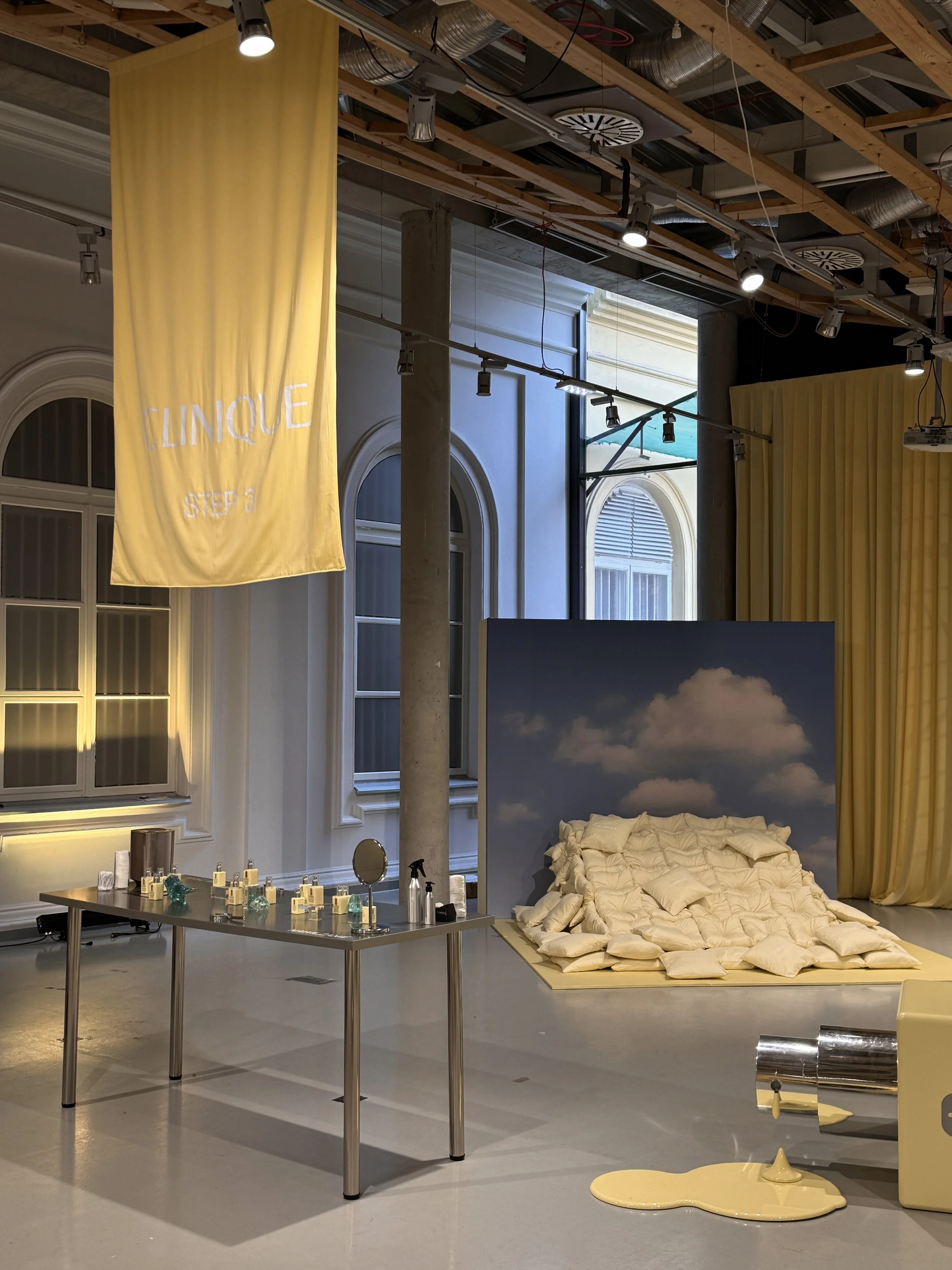 An art installation or display in an industrial-style room showing a bed with crumpled beige fabric, a table with bottles and objects, a yellow banner with white text that reads 'CLINIQUE STEP 3', and a yellow melting object on the floor