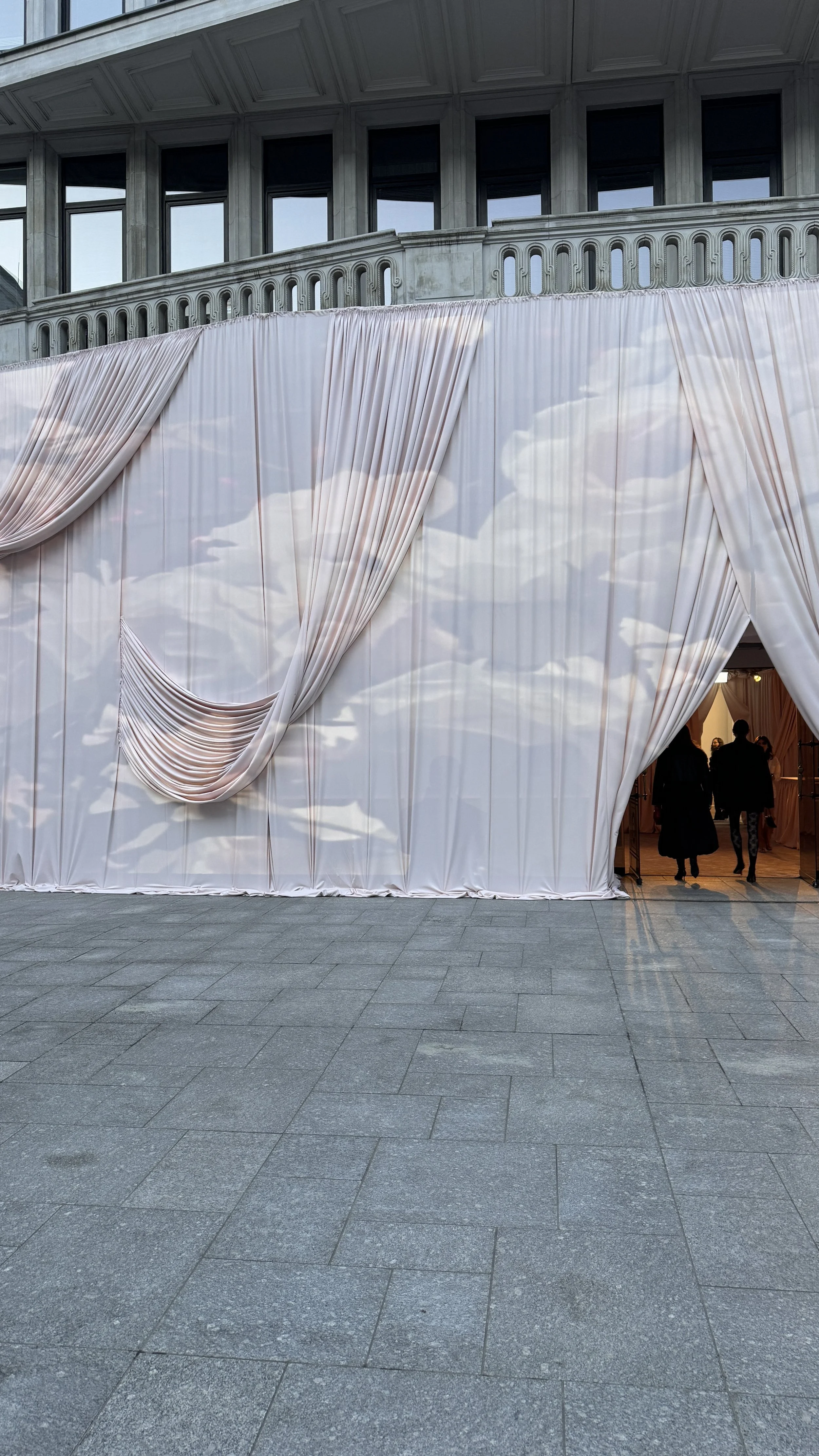 Large white curtain with draped fabric and cloud reflections, forming an entrance to a building, with two people walking inside.