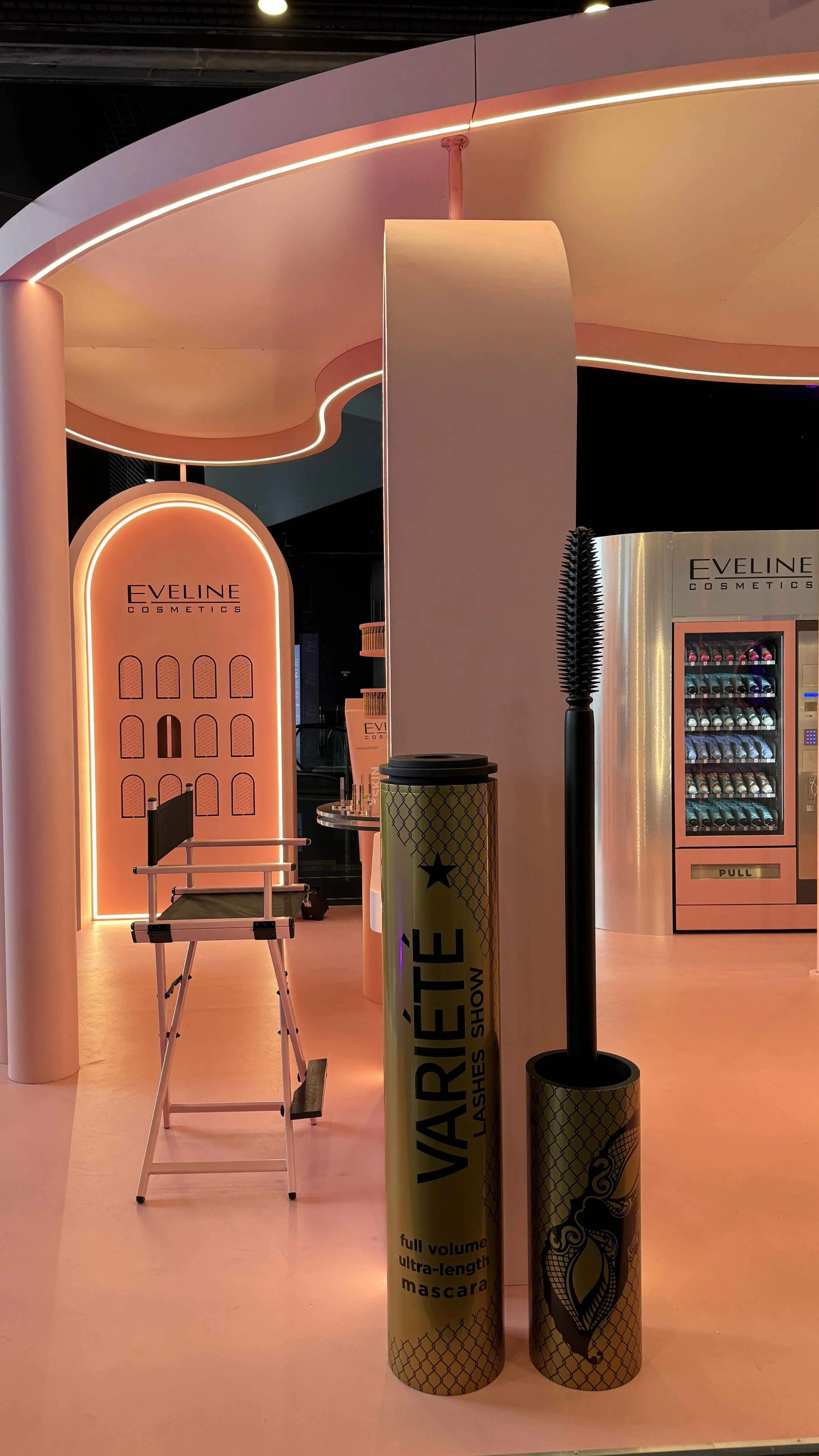 Display of cosmetics and beauty products at a store, featuring a vintage-inspired makeup chair, a vending machine with cosmetics, and large tube-shaped packaging labeled 'VARETTE' with full volume ultra-length mascara.