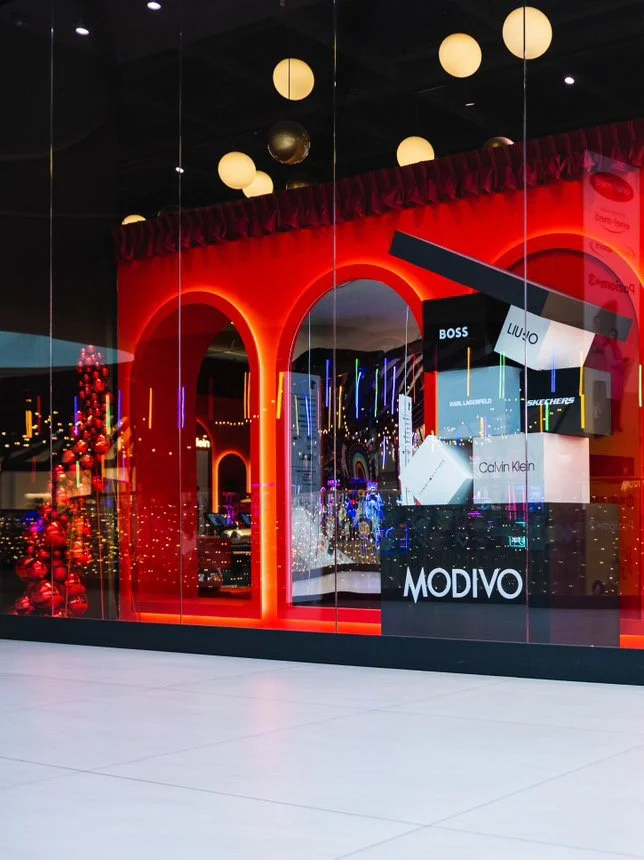 Store window display with multiple brand signs including MODIVO, Calvin Klein, BOSS, LIU JO, and SKETCHERS. Inside, festive Christmas decorations and lights are visible, with a Christmas tree decorated with red ornaments to the left.